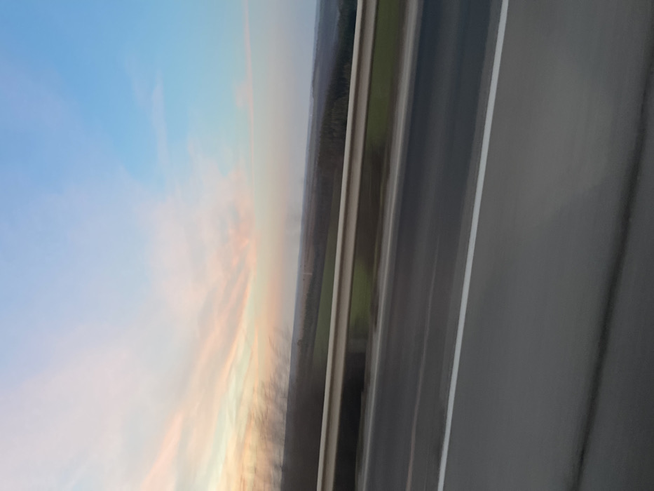 A panoramic landscape image captured from a moving vehicle in the vicinity of Osenets, Bulgaria, likely during dusk or dawn, is presented. The foreground features a grey asphalt road with a solid white lane marking on the right edge, overlaid by a light-colored metal highway guardrail, both exhibiting significant motion blur. Blurred dark vegetation is present on the extreme left. The mid-ground displays an expansive, undulating terrain composed of green agricultural fields and dense, dark forested areas. The background is dominated by a sky gradient, transitioning from soft pink and orange wispy clouds on the left to a clear light blue on the right. Faint, dark plumes or haze are discernible on the distant horizon. No individuals or other specific objects are identifiable.