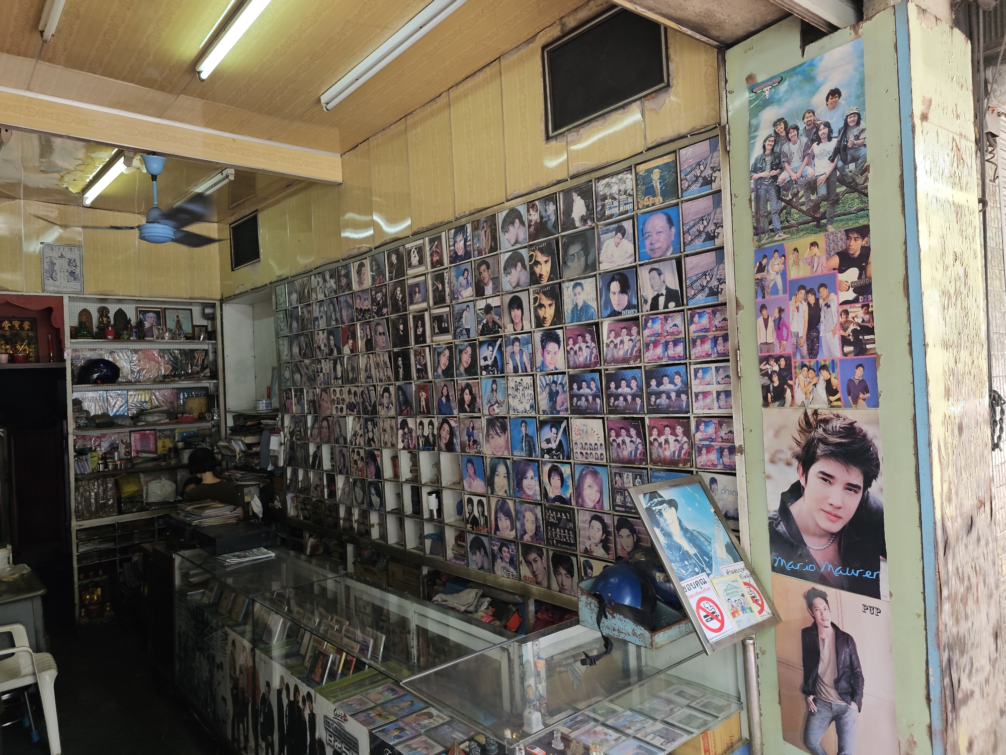 The image depicts the interior of a small, cluttered shop in Bangkok, Thailand, likely a music or entertainment store. The walls are extensively decorated with numerous posters and framed pictures, primarily of Asian musicians and celebrities, arranged in a grid-like fashion. Many of these images appear to be album covers or promotional photos.
In the foreground, there are glass display cases filled with what appear to be CDs or other media. Behind these cases, a counter is visible, stacked with more merchandise and items, including what looks like a blue helmet. A person is partially visible behind the counter, seemingly engaged with the merchandise.
The shop interior is somewhat dimly lit, with fluorescent lights on the ceiling. The walls are covered in a pale, textured material, possibly wood paneling, and show signs of wear and tear, with peeling paint and visible repairs. A blue ceiling fan is visible near the ceiling in the upper left portion of the image. The overall impression is of a densely packed, perhaps older establishment, catering to a specific niche market.