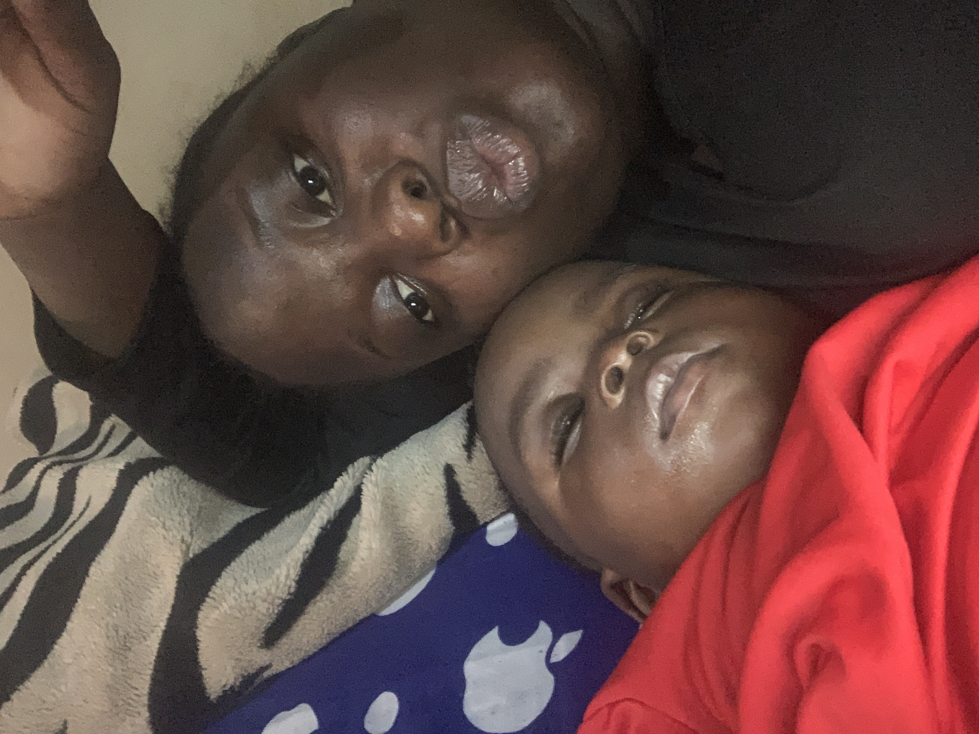 An overhead, close-up selfie shows a dark-skinned adult and a dark-skinned baby lying down, side-by-side. The adult's face is in the foreground, with their lips pursed, and one hand is raised above their head. The baby's face is to the left of the adult's, with their eyes closed. Both are wearing dark clothing. Behind them, a blanket with a black and white zebra stripe pattern is visible, as well as a blue fabric with white apple logos. The environment is dimly lit and appears to be a bedroom.