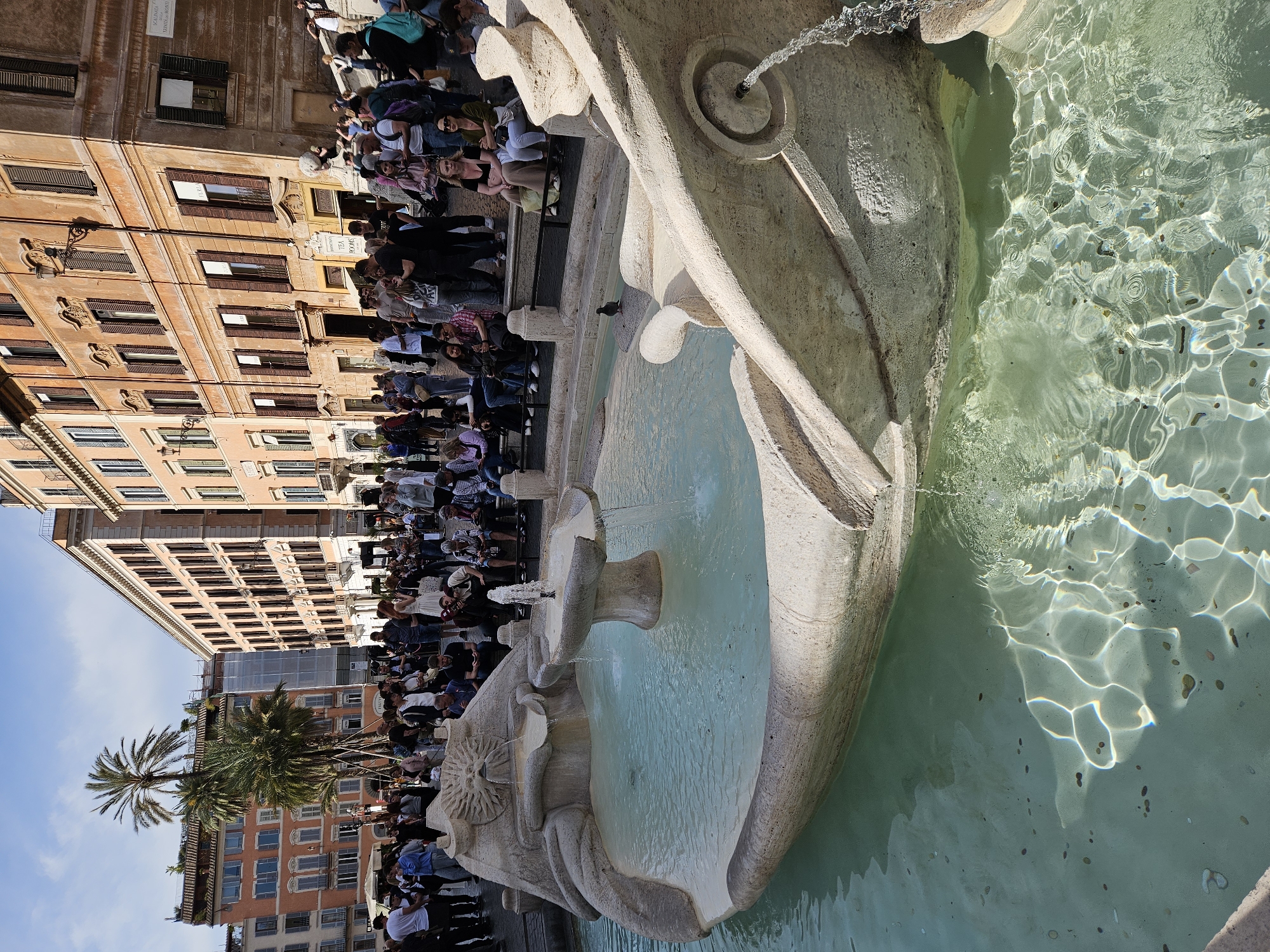 A horizontal photograph depicts the Fontana della Barcaccia, a baroque fountain in Piazza di Spagna, Rome, Italy. The fountain, made of white travertine, is positioned in the right half of the frame. Water flows from the fountain's elements, creating reflections and refractions on the water's surface. Numerous coins can be seen in the water. A group of people sits on the steps behind the fountain on the left side of the image. Buildings are visible behind the crowd, with a palm tree adding a touch of greenery. The sky is visible above the buildings.