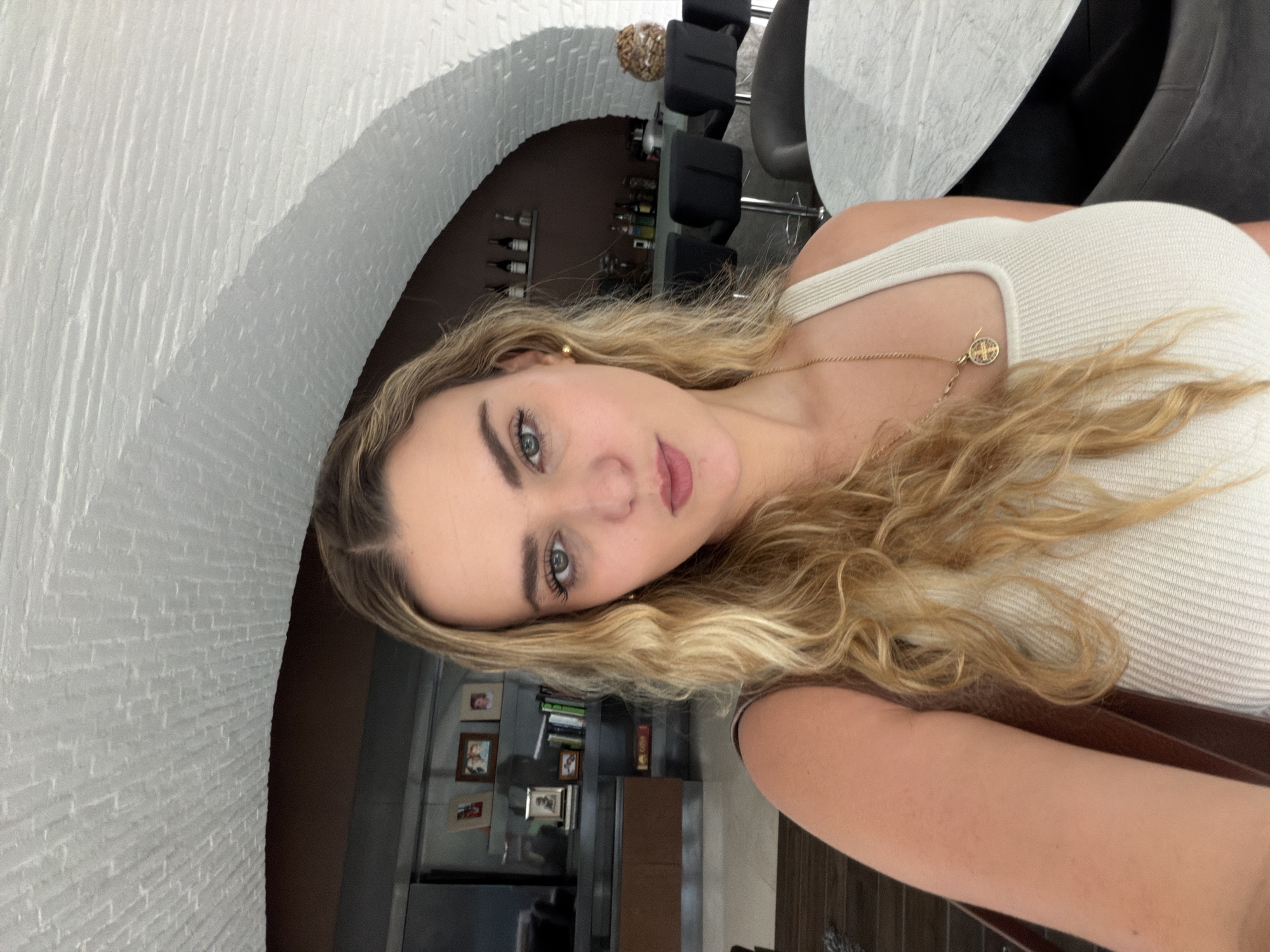 A young woman with long, wavy blonde hair and light-colored eyes takes a selfie in an indoor setting. She is positioned diagonally in the frame, wearing a light-colored, ribbed tank top and a delicate gold chain necklace with a small, round, multi-colored pendant. A small gold hoop earring is visible in her left ear. Her lips are slightly parted in a soft pout.
The background features a white brick archway. Through the archway, a darker room is visible with shelves holding numerous bottles, possibly wine or spirits. To the left of the woman's head, there's a bookshelf or display unit adorned with several framed pictures and books. To her right, parts of a modern table with a light-colored top (possibly marble) and dark chairs are visible. The overall setting appears to be an upscale interior space, possibly a home or a chic restaurant/lounge. There is no visible text in the image.
