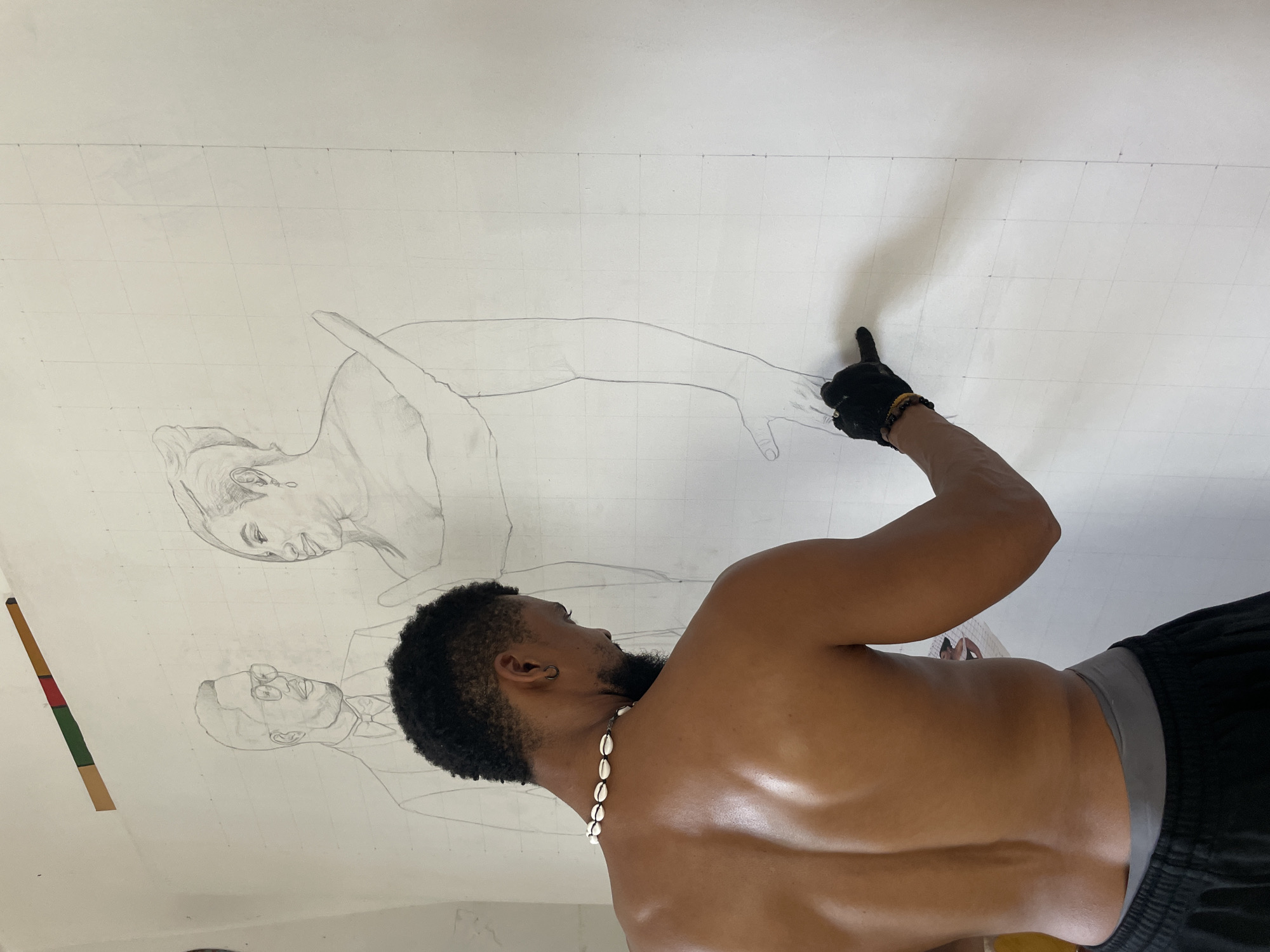 A torso of a dark-skinned man is captured in a close-up, angled upwards. He wears black shorts with a grey waistband and a necklace of white seashells. His black hair is styled with a short cut, and a dark beard is visible on his face. He points with his right index finger, which is covered with a black glove.
Above the man, a large white canvas is stretched out. The canvas has a grid pattern drawn on it and two pencil sketches are visible. The upper sketch shows a woman's face and upper body. Below her is a man's face wearing glasses, possibly a portrait.
A strip of tape with green, yellow, and red colors is visible on the far left of the canvas.
The image could be inside a studio in Ado, Nigeria. The location is not definitively verifiable from the given image.