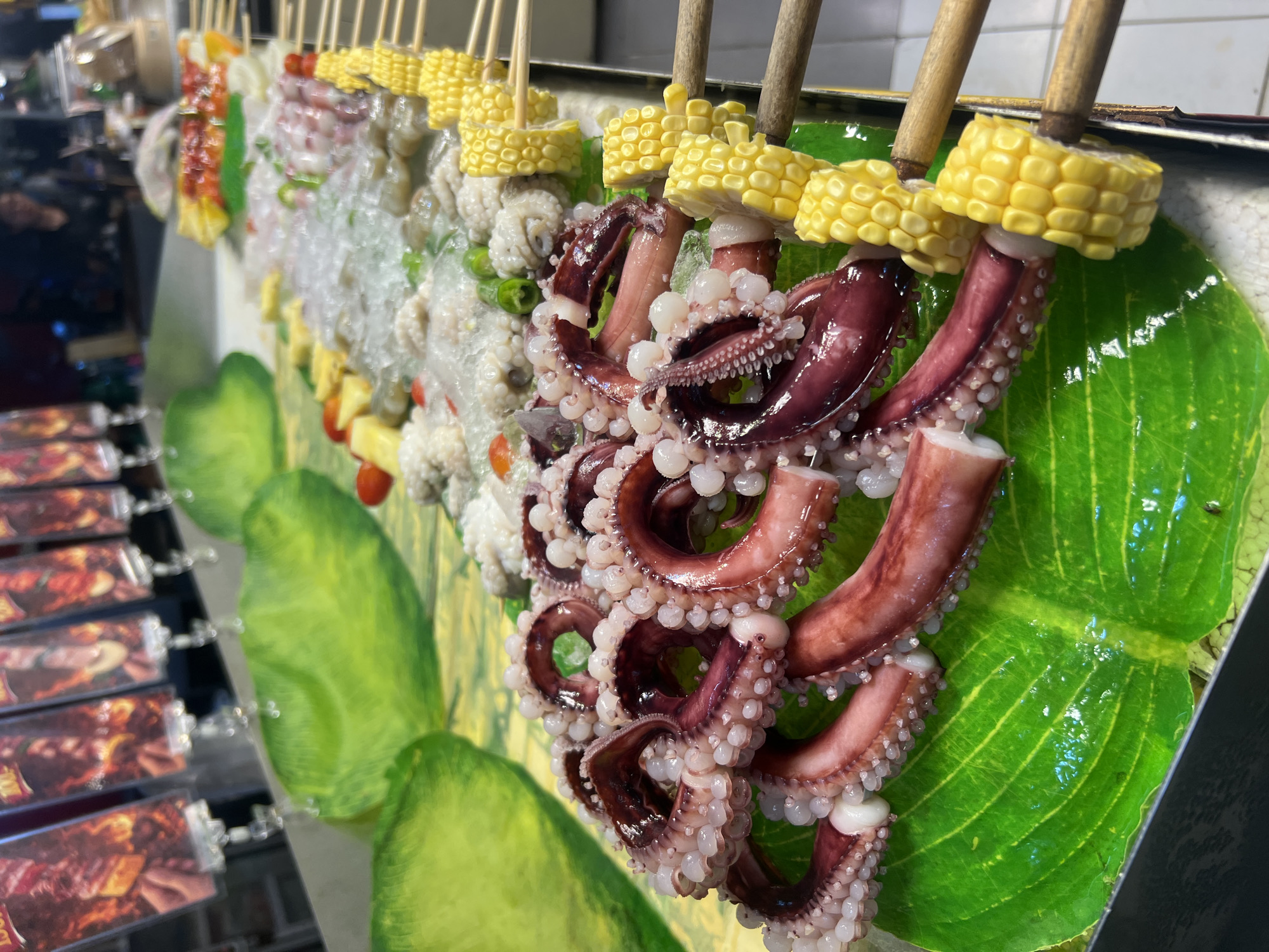 This image showcases a vibrant street food display in Karon, Thailand, focusing on a variety of seafood skewers. The most prominent subject is a set of grilled octopus tentacles, characterized by their deep reddish-purple color and an abundance of suction cups. These tentacles are skewered with pieces of bright yellow corn on the cob.
To the left of the octopus, other skewers are visible, including what appears to be small whole octopuses, some possibly covered in ice or a light coating. Further down the line, skewers with seafood and other ingredients like cherry tomatoes and pineapple chunks are partially visible, along with some green vegetables, possibly peppers. The food is arranged on a bed of decorative green leaves, which add to the visual appeal of the stall. In the background, the setting suggests a bustling market or street food area with other stalls and displays, though these are out of focus.
The lighting indicates it could be daytime or early evening, typical for street food operations. The overall impression is one of fresh, exotic food presented attractively to potential customers. There are no people clearly visible in the foreground, and no text is legible in the image. The focus is entirely on the appetizing seafood skewers, implying a culinary experience being offered.