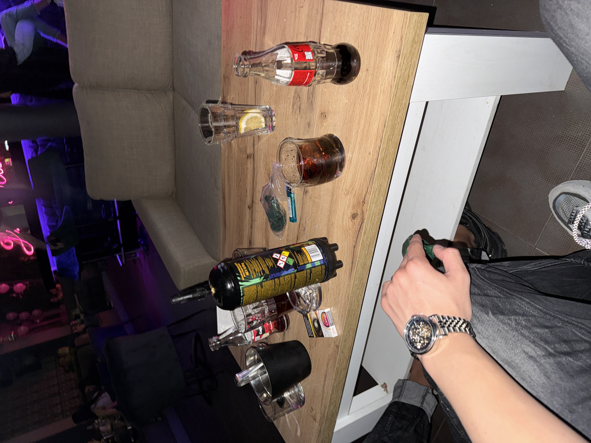 The image shows a scene on a table, likely in a bar or nightclub setting. The table is covered with various objects, including empty liquor bottles (Coca-Cola, Jagermeister), glasses, and a gas canister. The setting suggests a social gathering where alcohol consumption is taking place.
A person is sitting near the table, as indicated by their hand and wristwatch being visible. Their attire includes a denim jacket, indicating a casual setting.
The location is identified as Karlovo, Bulgaria.