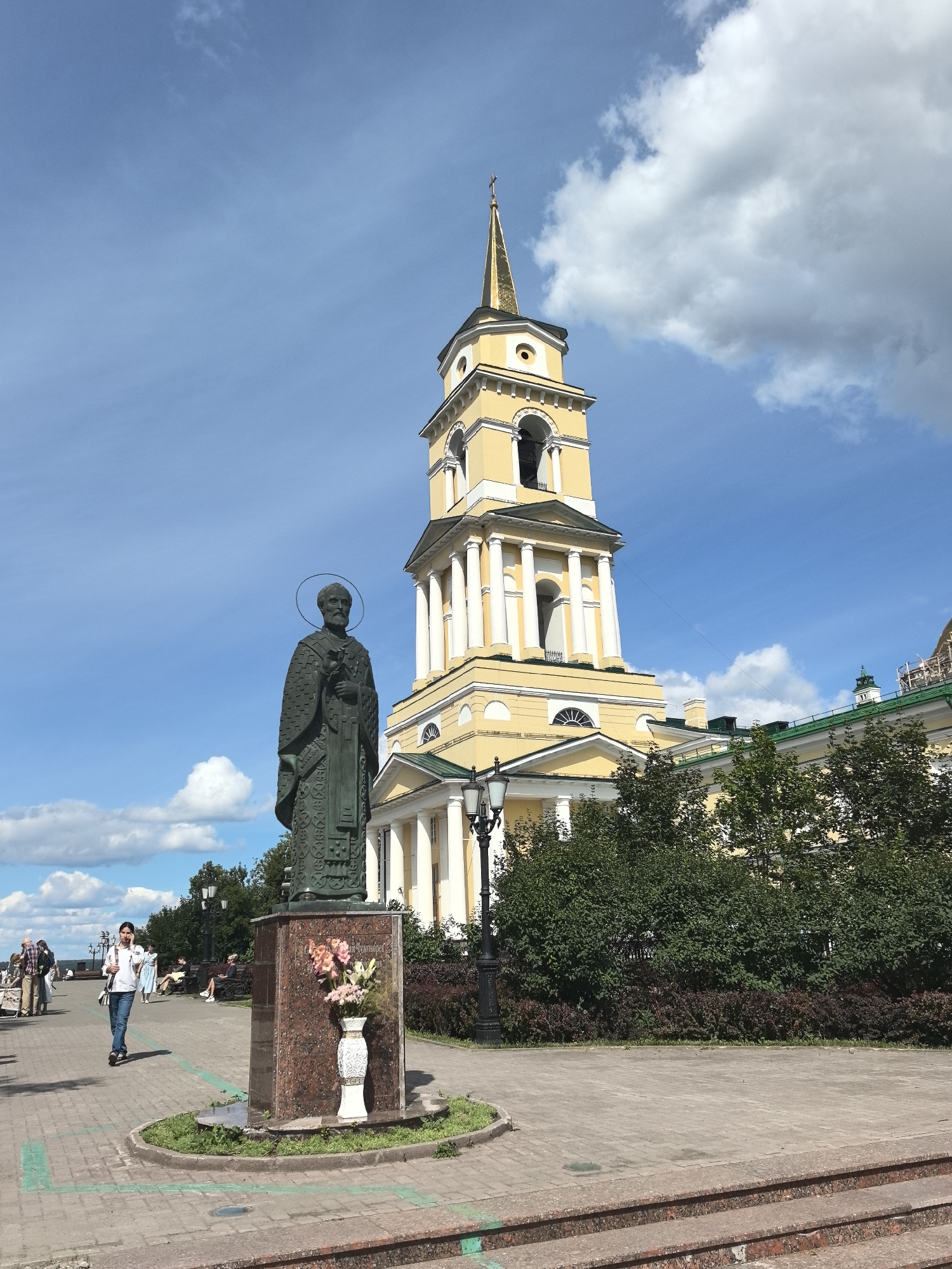 The image depicts a bronze statue of a man with a halo in the foreground, standing on a granite pedestal. The statue is located in a park-like setting with paved walkways, benches, and lush green trees and bushes. In the background, a large yellow building with white columns and a tall bell tower with a golden spire is visible. A few people are scattered throughout the scene, some walking and others sitting on benches. One person is seen walking in the foreground near the statue, while others are further in the distance. The sky is blue with scattered white clouds, suggesting fair weather. The overall impression is a serene and picturesque urban landscape.
This scene is located in the city of Perm, Russia, specifically in the Perm State Art Gallery, where the monument to the physician and philanthropist Fedor P. Belikov stands. The building in the background is indeed the former Cathedral of the Transfiguration, which now houses the Perm State Art Gallery.
