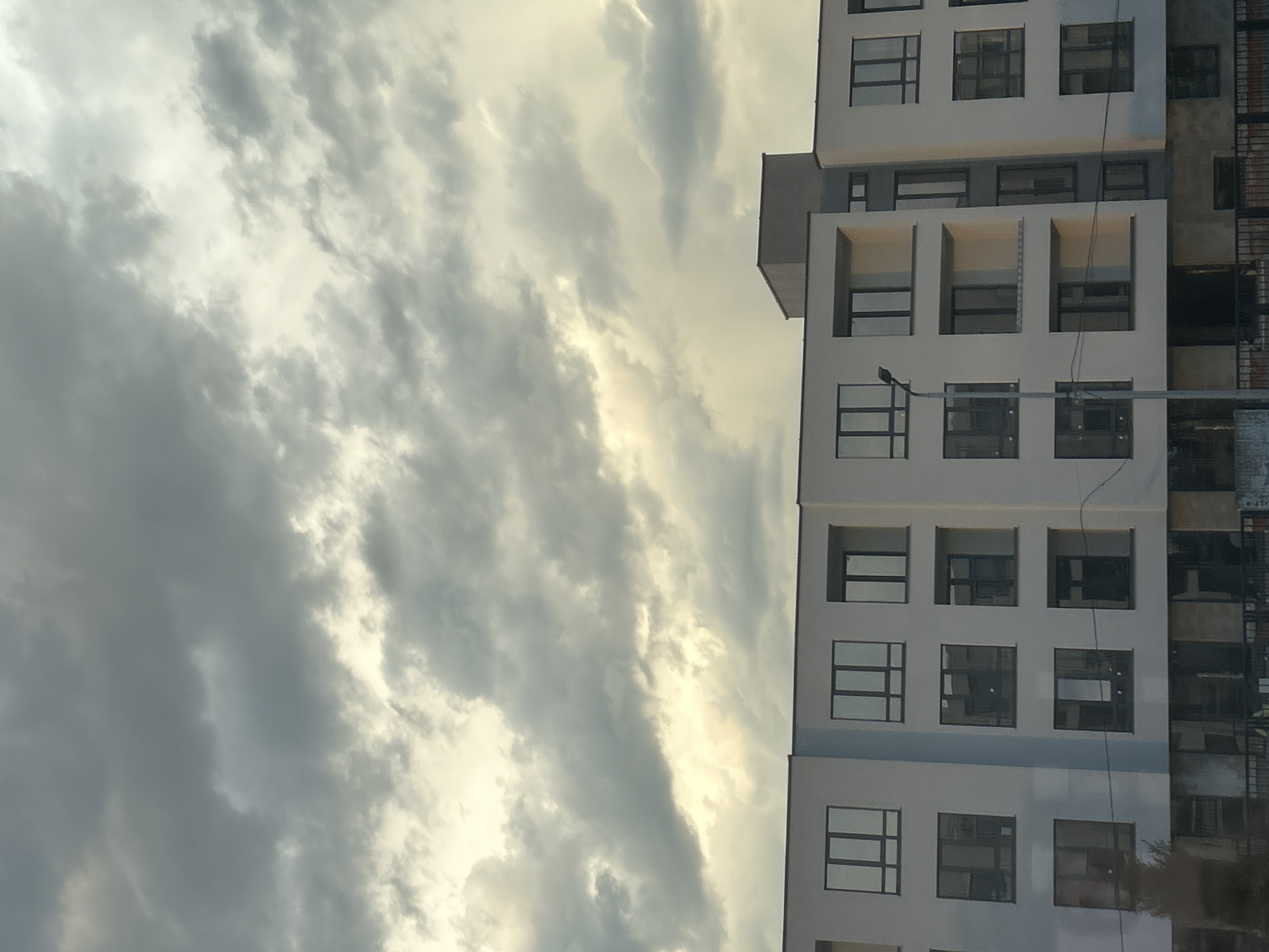 The image shows a multi-story building in Plovdiv, Bulgaria, viewed from a low angle with a cloudy sky in the background. The building has a white facade with multiple rows of windows. Some of the windows have protruding balcony-like structures. To the right of the building appears to be a construction site or unfinished section, with scaffolding or metal structures visible. The sky is filled with dense, gray clouds with patches of brighter light breaking through. A utility pole with a single street lamp extends upwards in front of the building. A few power lines run across the face of the building. The overall impression is that of a somewhat overcast day.