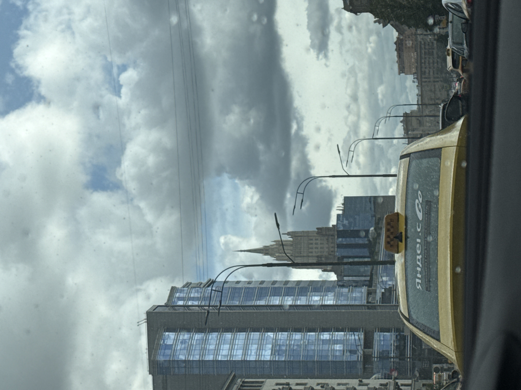 The image captures a street scene in Moscow, Russia, likely taken from inside a vehicle, with a yellow Yandex taxi in the foreground. A modern glass office building is seen in the mid-ground, and the Ministry of Foreign Affairs building is visible in the background. The sky is overcast with varying shades of gray and white clouds. Streetlights and power lines are visible above the buildings, contributing to the urban environment. There are other cars visible on the road as well.