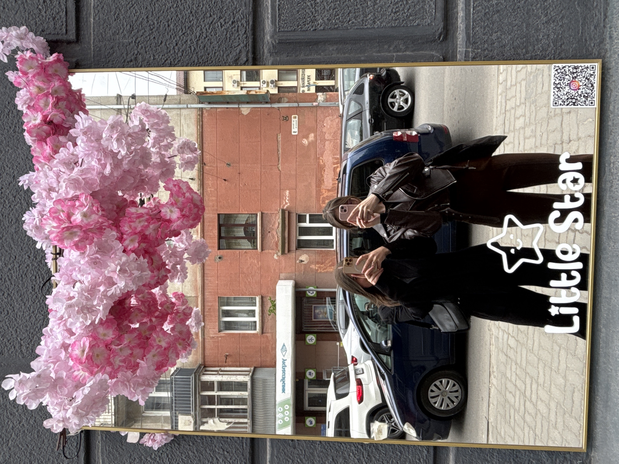 A mirror is reflecting a street scene with two women, cars, and buildings. The mirror has a gold frame and is decorated with pink flowers on the left side, and white text reading "Little Star" on the right, along with a QR code. The women are positioned in front of the mirror, seemingly taking a selfie. The woman closer to the camera is wearing a brown leather jacket and brown pants, while the other woman is wearing a black outfit. Cars are parked along the street, including a dark blue car and a white car. The buildings across the street are made of red brick and white concrete. A sign on the building reads "Industriabank" in a Cyrillic script. The location is a city of Unknown, Unknown.