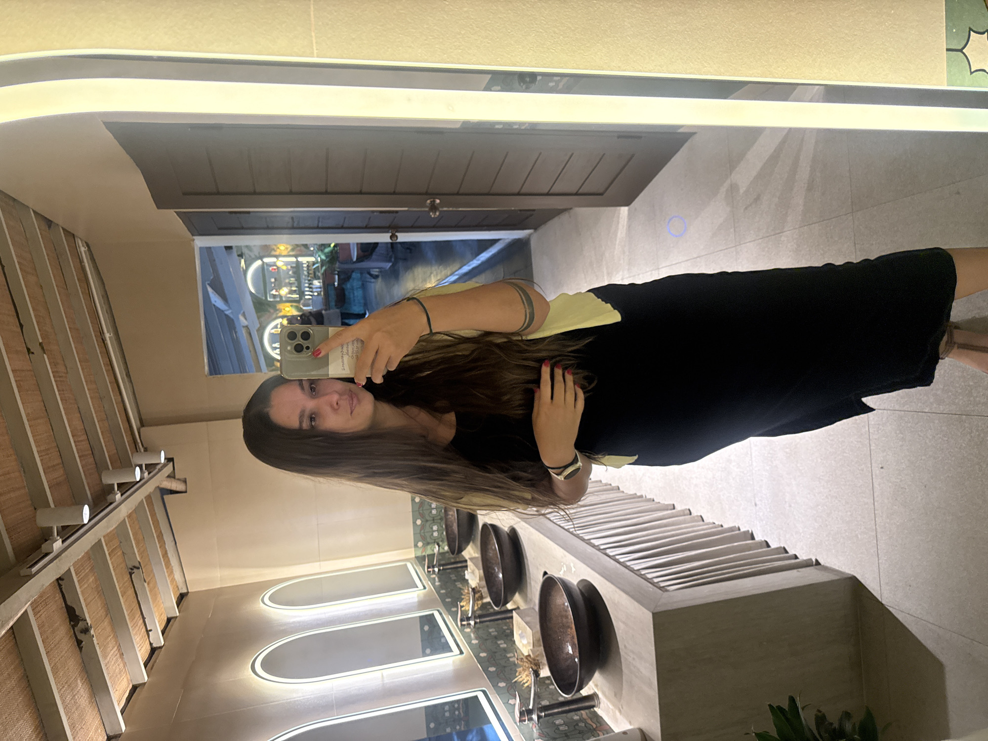 A full-body selfie of a young woman with long brown hair is taken in a modern bathroom. She is wearing a black sleeveless dress and a light yellow cardigan. The woman is holding a smartphone in her right hand, angled to capture her reflection in a large, illuminated mirror. The bathroom features a long countertop with several dark, bowl-shaped sinks and faucets. Above the sinks, a row of arched mirrors is illuminated from within. The ceiling is made of woven material supported by white beams, with track lighting visible. In the background, through an open doorway, a glimpse of a brightly lit interior with a bar area can be seen. The floor is tiled with light-colored square tiles. The overall impression is of a stylish and contemporary restroom.