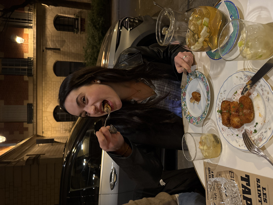 The image depicts an evening outdoor dining scene on a white circular table, likely a café terrace in Paris, France. The menu visible on the table explicitly references "TAPAS LES SAINT" and "4 RUE DES CAVES," an address located in Paris.
A young woman with long dark hair, wearing a black leather jacket over a plaid shirt, is the primary subject. She is smiling with her mouth open, actively bringing a forkful of food to her mouth from a small plate containing a single breaded item, possibly a croquette. She wears a pearl earring on her right ear, a ring on her left ring finger, and a necklace with a 'G' pendant. Partially visible to the left is another person's arm, suggesting shared dining.
On the table, another plate holds three similar breaded and fried items, garnished with red spice and green herbs, alongside a fork and knife. A clear glass pitcher filled with an amber liquid, ice, and fruit slices is present, along with two individual glasses containing the same beverage. A clear patterned glass ashtray rests on the menu card.
The background reveals a paved street with a parked silver SUV. Behind the vehicle stands a classic Parisian building of light-colored stone, featuring arched ground-floor windows, rectangular upper-story windows, and wall-mounted lanterns, all illuminated by warm artificial light, indicating nighttime.