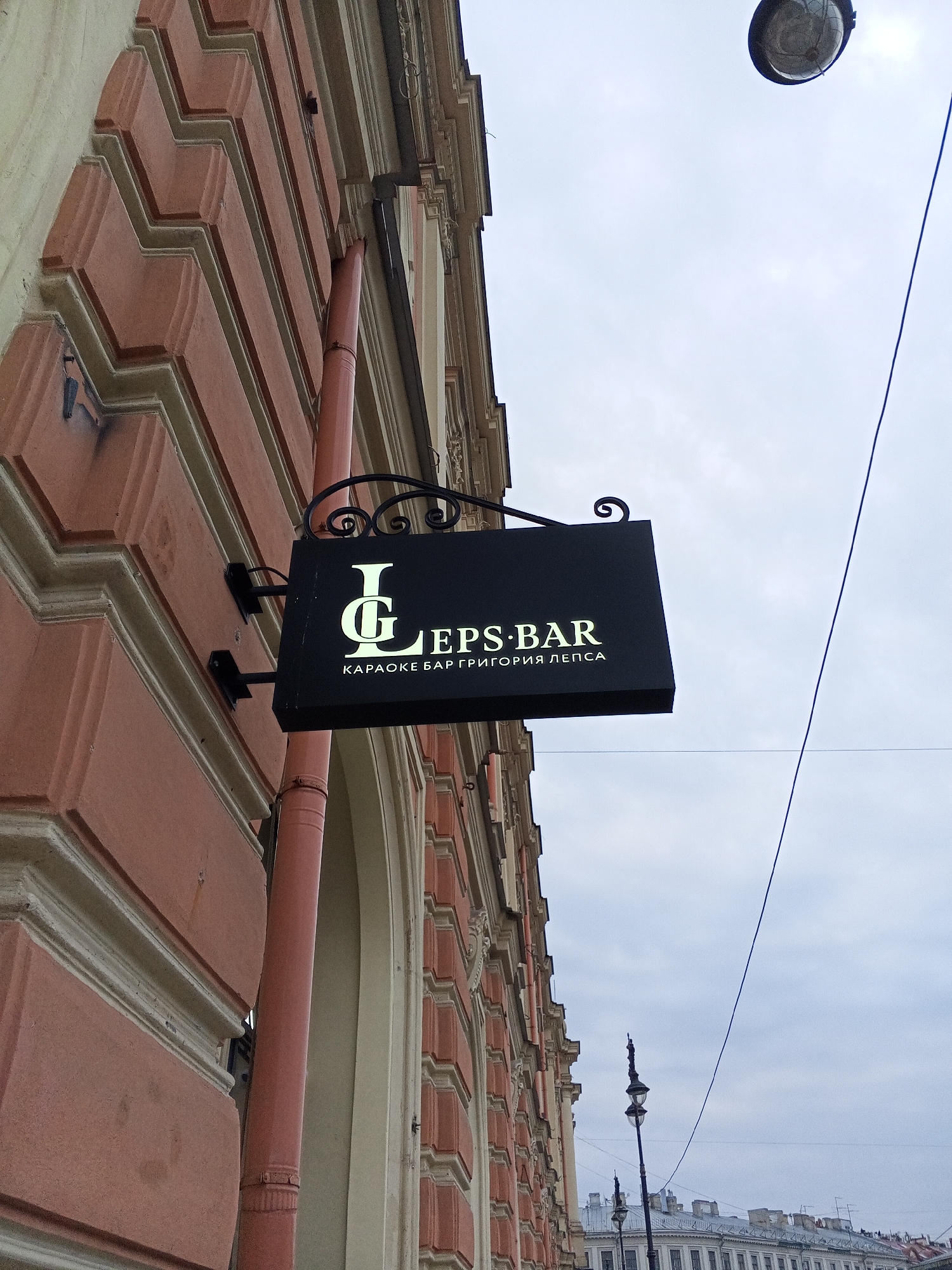 The image depicts a street scene in Saint Petersburg, Russia, featuring the sign of "LEPS BAR karaoke bar Grigoriy Lepsa" attached to the side of a building. The building is ornate, with pink and beige architectural details, and a red drainage pipe is visible running along the side. Above the sign, a street lamp is partially visible against a cloudy sky. In the background, other buildings are seen. A thin black wire runs diagonally across the sky. The scene appears to be during the daytime, under overcast weather conditions.