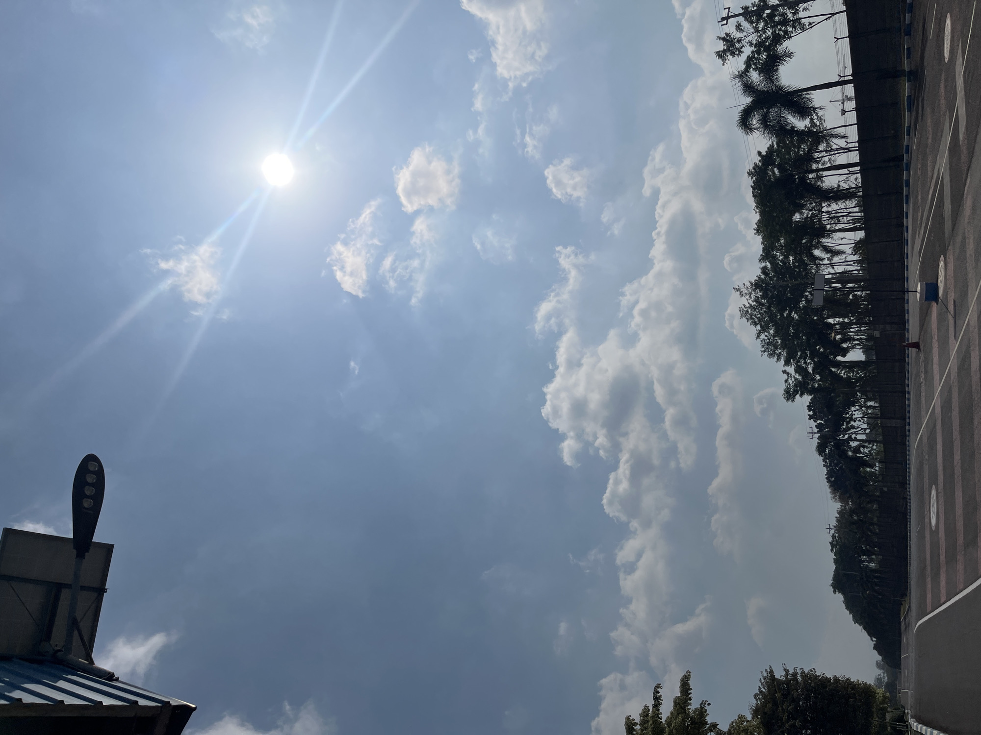 The image depicts an outdoor scene, likely in a suburban or industrial area of Pelang, Indonesia, on a bright, sunny day. The sky is a clear blue, dotted with fluffy white clouds. The sun is high in the sky, casting bright rays that create a lens flare effect. In the foreground, there is an asphalt road with white lane markings and what appear to be parking lot dividers. To the right of the road, there is a dark fence with a blue and white striped border, behind which are rows of various trees, including palm trees. In the distance, a construction crane is visible. To the left of the road, there are more trees. In the upper left corner of the image, a modern street lamp with multiple lights is mounted on a structure that appears to be a roof or awning. There are no people visible in the image.