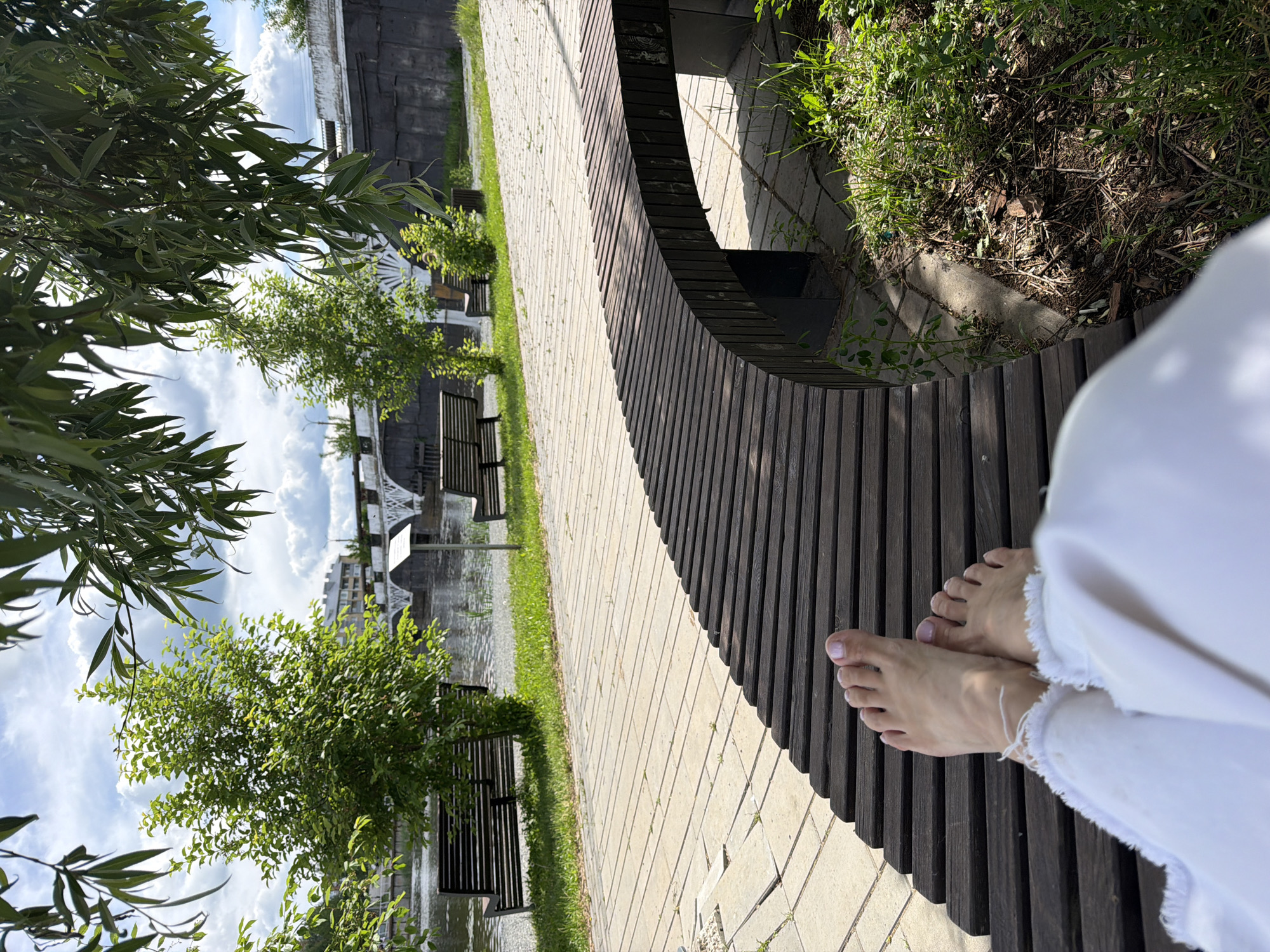 The image captures a first-person perspective of someone relaxing outdoors in what appears to be a park or riverside area in Votkinsk, Russia, during daytime. The primary subject is a pair of bare feet, presumably belonging to the photographer, resting on a curved, slatted wooden bench. The feet show a light-colored nail polish on the toes, and are partially covered by the frayed hem of white pants or trousers.
The setting features a paved pathway made of light-colored rectangular tiles, which runs alongside the wooden bench. Beyond the path, there is a strip of green grass and lush foliage, including several small, bushy trees. In the foreground on the left, a leafy branch with long, slender, willow-like leaves hangs into the frame.
In the mid-ground, a body of water, likely a river or canal, is visible, reflecting the cloudy sky. Across the water, there are structures that suggest a bridge or industrial buildings, partially obscured by trees. The sky is bright with white cumulus clouds. Sunlight casts clear shadows on the bench and pathway, indicating a sunny day. No legible text is visible in the image.