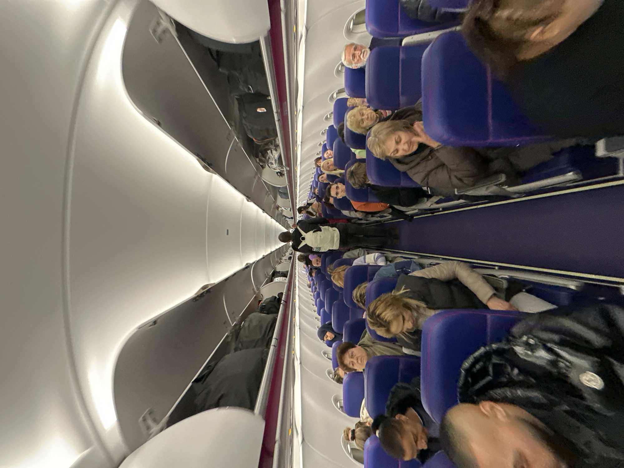 The image shows the interior of an airplane cabin. Passengers are seated in rows of blue seats, some with their heads reclined. An aisle runs down the center of the cabin. Overhead bins line the top of the cabin walls. An individual wearing a white shirt and backpack stands in the aisle. The overall environment is typical of an airplane interior. The city of Sofia, Bulgaria is not evident in this image.
