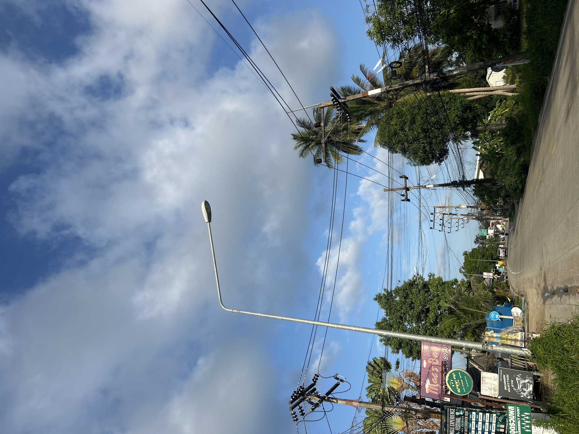 This is a street-level, eye-level view looking down a paved road in the Ko Pha-ngan District of Thailand. The sky is blue with scattered white and gray clouds. On the left side of the road, there are numerous signs, including one for "Macs Bay Resort" and another advertising a "Jungle Experience Festival." There are also palm trees, utility poles with many electrical wires, and lush green vegetation. The road curves to the right and continues into the distance, with more buildings and trees visible on either side. A few vehicles are present, including a white SUV and a motorcycle, as well as a single pedestrian walking in the distance. A street lamp is prominent in the foreground on the left.
