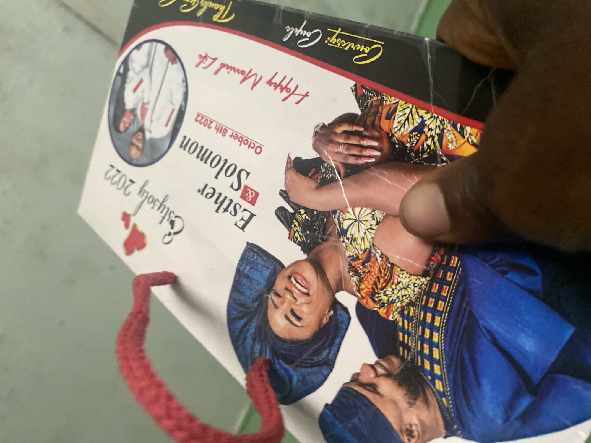 Here is a detailed description of the image:
The image shows a gift bag featuring a wedding announcement for Esther and Solomon, whose wedding took place on October 8th, 2022. The bag is held by a dark-skinned hand. The bag itself is white with a red handle. The wedding announcement includes the text "Esther & Solomon" and "Happy Married Life." There are also images of the couple. One image is a headshot of them dressed in traditional Nigerian attire: the woman is wearing a blue headwrap and the man is wearing a matching hat and outfit. Another image shows them in Western clothing. At the top of the bag, the text reads "Courtesy: Couple." There is also a mention of "Thanks for..." which is partially cut off. The location is noted as being in the city of Unknown, Unknown.