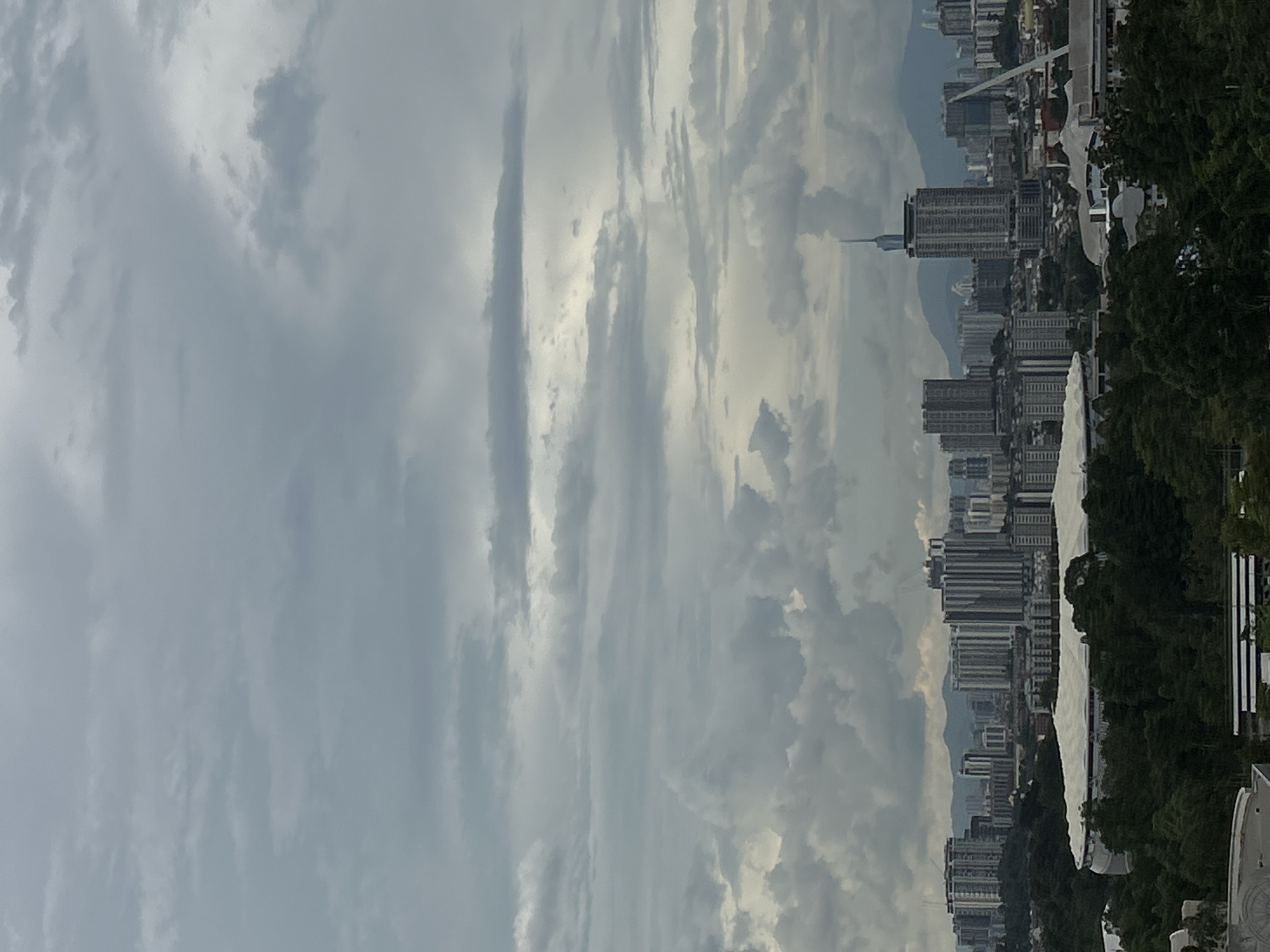 The image shows an urban landscape under a cloudy sky. In the foreground are tree tops. A cluster of buildings in the mid-ground suggest a city skyline, and a tall, thin spire is visible among the structures. The sky is predominantly grey with diverse cloud formations. The scene suggests Kuala Lumpur, Malaysia, based on the architectural style of the buildings, the presence of the spire (likely the KL Tower), and the tropical foliage.