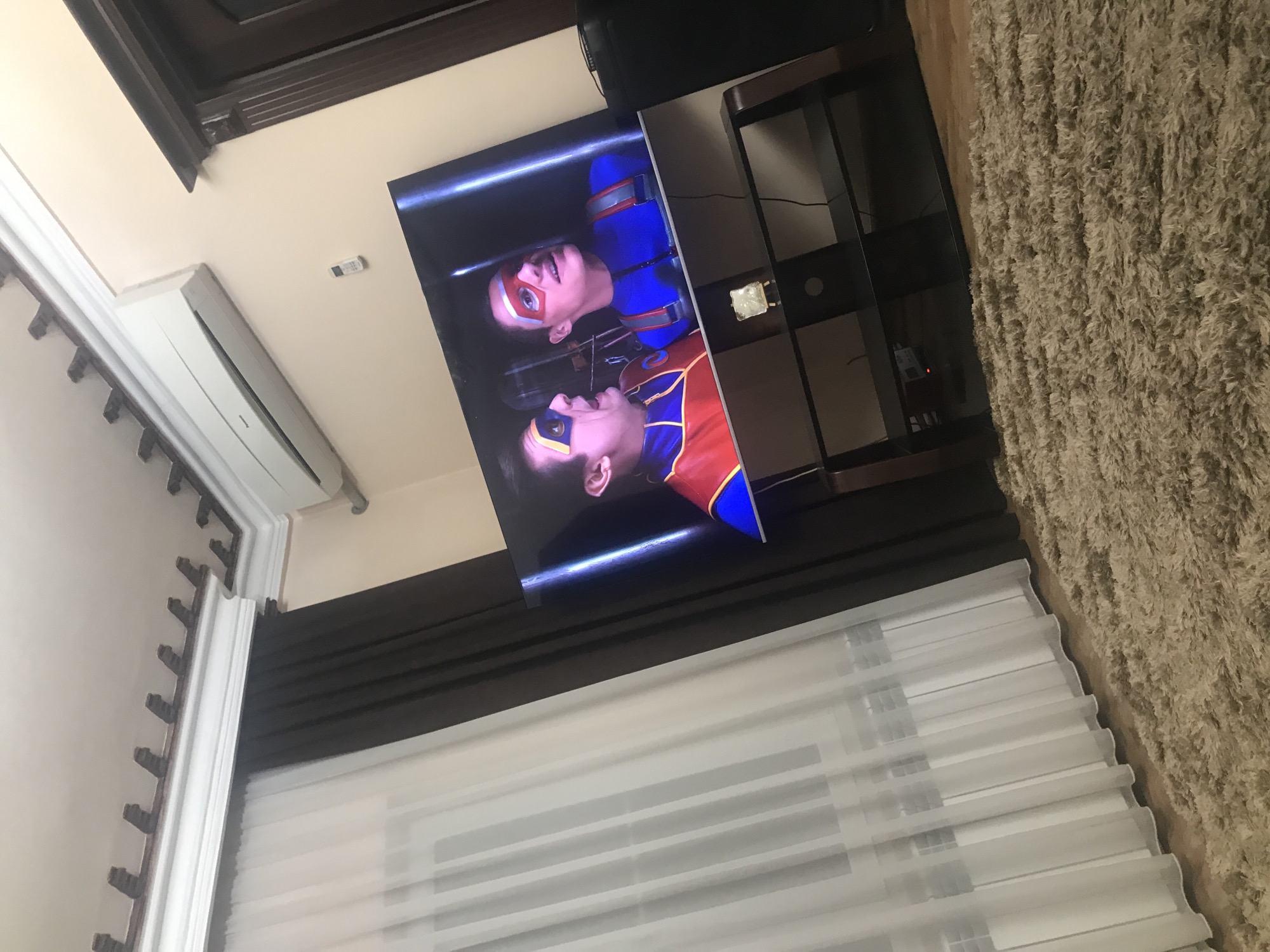 The image depicts a living room interior, possibly in Yerevan, Armenia, although there are no explicit identifiers confirming this. The view is angled, looking towards a television displaying characters in superhero costumes. Below the TV, there's a window covered with partially translucent white blinds, and adjacent to the TV stand, there's a shaggy, light brown rug. An air conditioning unit is mounted on the wall above the window. The ceiling has dark trim.