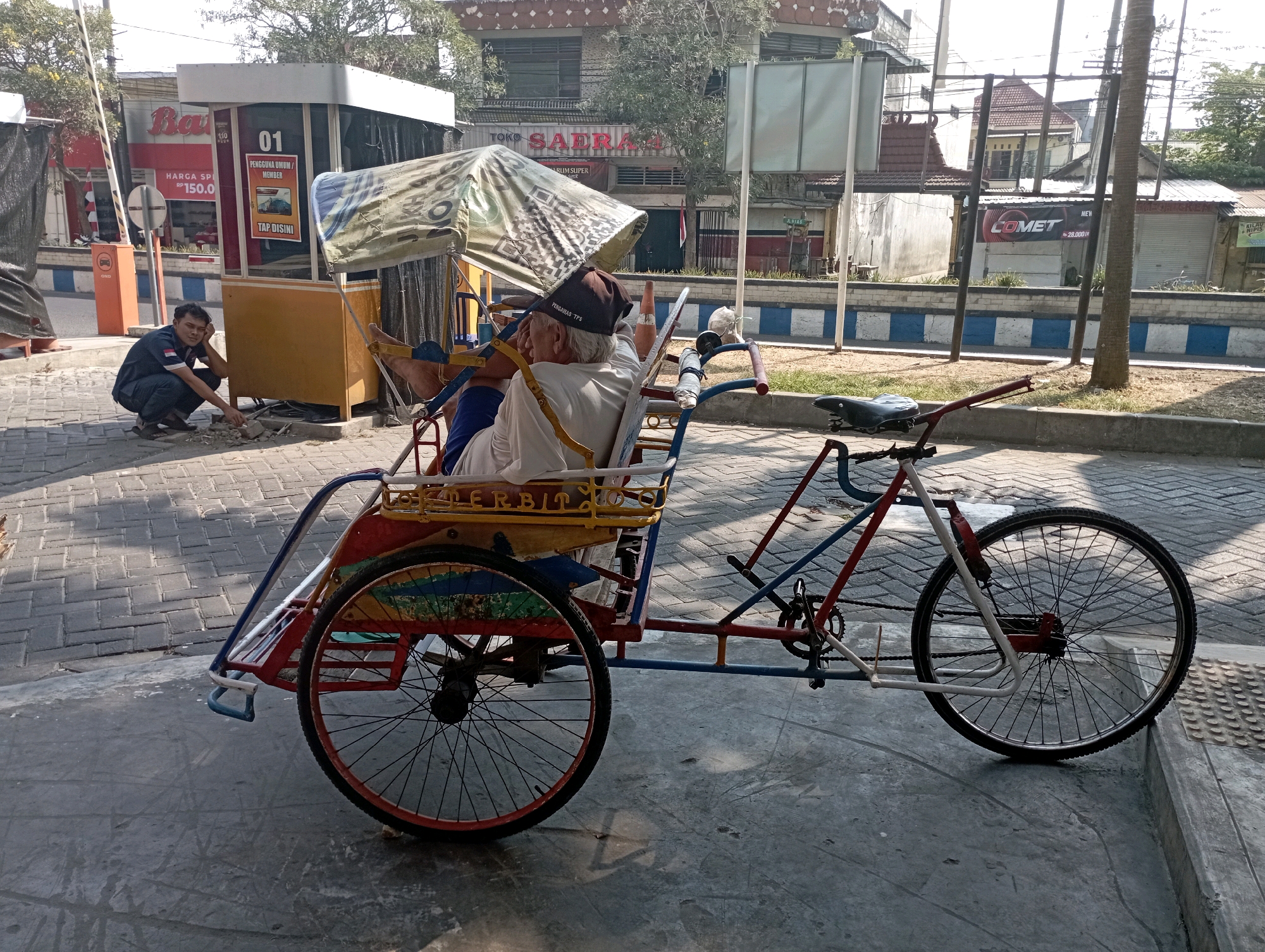 In the city of Ponorogo, Indonesia, a man is crouched down on a paved sidewalk, appearing to be attending to something on the ground near a small, yellow booth. The booth has a sign that says "01" and "PENGGUNA UMUM/MEMBER". Further back, a pedicab, a three-wheeled vehicle powered by a person pedaling, is parked. An elderly man wearing a hat is seated in the passenger section of the pedicab, under a patterned canopy. The pedicab has a colorful frame with red, blue, and yellow accents and spoked wheels. Behind the pedicab and booth, buildings and trees are visible, indicating an urban setting. The scene is illuminated by sunlight, casting shadows on the ground.
