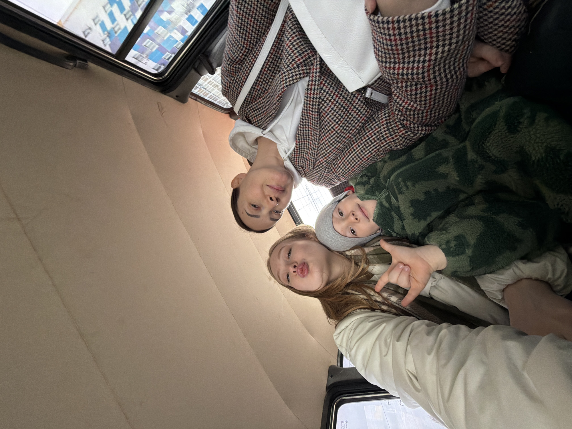 A low-angle photograph, rotated counter-clockwise, captures an adult woman, a teenage girl, and a young child inside a vehicle, likely a car or van, in Засечное, Russia. The shot appears to be a group selfie or portrait taken from below, looking up at the subjects and the vehicle's ceiling.
In the composition (when rotated upright), an adult woman is visible in the upper left, looking directly at the camera with a neutral expression. She wears a patterned tweed or houndstooth jacket over a white hoodie and carries a white shoulder bag. To her right and slightly below is a teenage girl with long, light-colored hair, looking at the camera with pursed lips, making a playful "duck face." Her right hand is raised, forming a "rock on" gesture. She is dressed in a light-colored jacket or coat. To the girl's right is a young child, whose face is partially visible, also looking towards the camera. The child wears a grey beanie and a green camouflage-patterned fuzzy jacket.
The setting is the interior of a vehicle, characterized by a light beige or tan upholstered ceiling, which appears to have a padded or quilted texture. Through the windows in the background, reflections of modern residential buildings with blue and white facades are visible, indicating an urban or suburban environment. The lighting suggests it is daytime, possibly overcast, given the soft and even illumination. The overall mood conveyed by the subjects' expressions and poses is casual and playful. There is no visible text in the image.