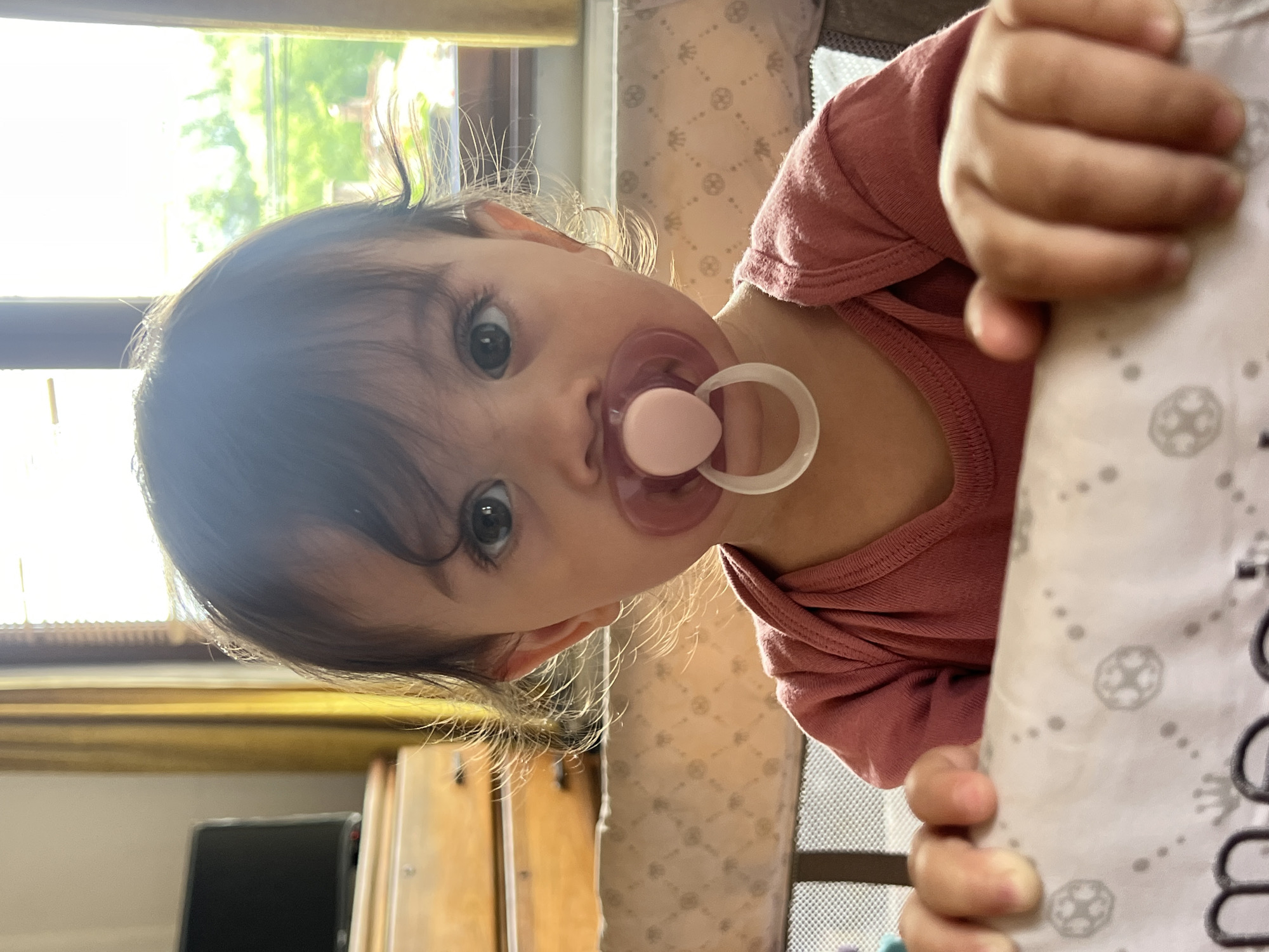 A baby with a pacifier in their mouth is shown in a close-up shot. The baby is wearing a pink shirt and is holding onto a white fabric with a gray pattern. The background includes a window, a wooden dresser, and a glimpse of greenery outside. The location is Elena, Bulgaria, but there is no information that verifies it is the city of Elena.