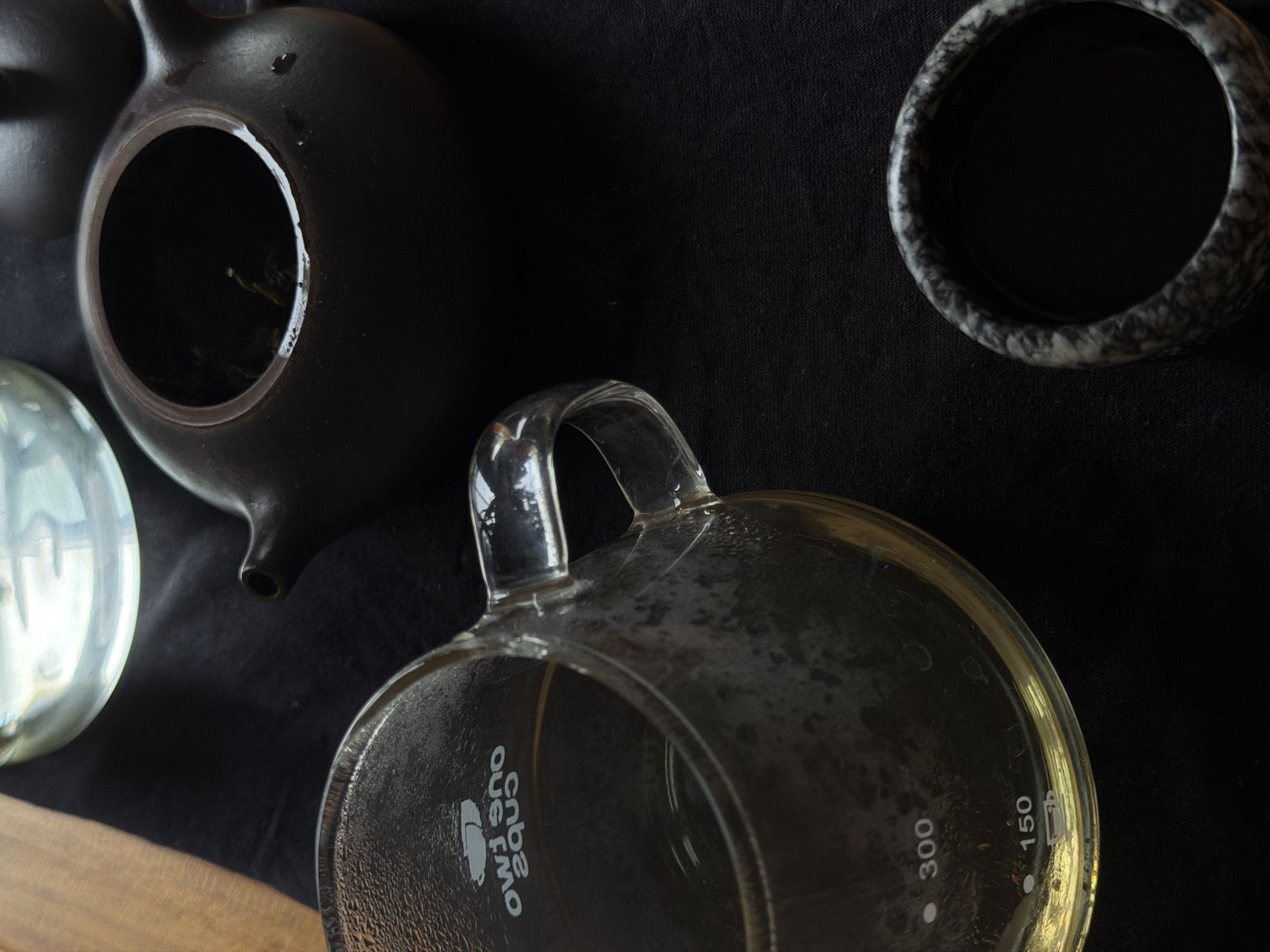 The image displays a still life composition of tea-related items arranged on a black fabric surface, with a wooden surface partially visible on the lower left. The perspective is an overhead, slightly tilted shot.
In the upper left quadrant, a dark brown, matte-finished ceramic or clay teapot is featured. Its lid is removed, revealing an open circular top with dark green tea leaves visible inside, along with some moisture droplets around the inner rim.
Centered in the frame and lying on its side is a clear glass measuring cup or mug with a prominent handle. The cup contains a light yellow, translucent liquid, likely brewed tea. Visible on its exterior are some water marks or residue. Mirrored and inverted text, "cnbs oub smo," is etched on the side of the glass cup, accompanied by a stylized mug logo. Measurement markings "300" and "150" are also present inside the cup.
In the upper right, a dark grey or black teacup with a marbled or speckled pattern is partially visible. This cup also appears to contain a dark liquid. A clear, spherical glass object reflects light on the far left edge of the frame.
The arrangement suggests a moment during tea preparation or serving. The scene is set in North Kuta, Indonesia, with controlled indoor lighting highlighting the textures of the various objects against the dark background.