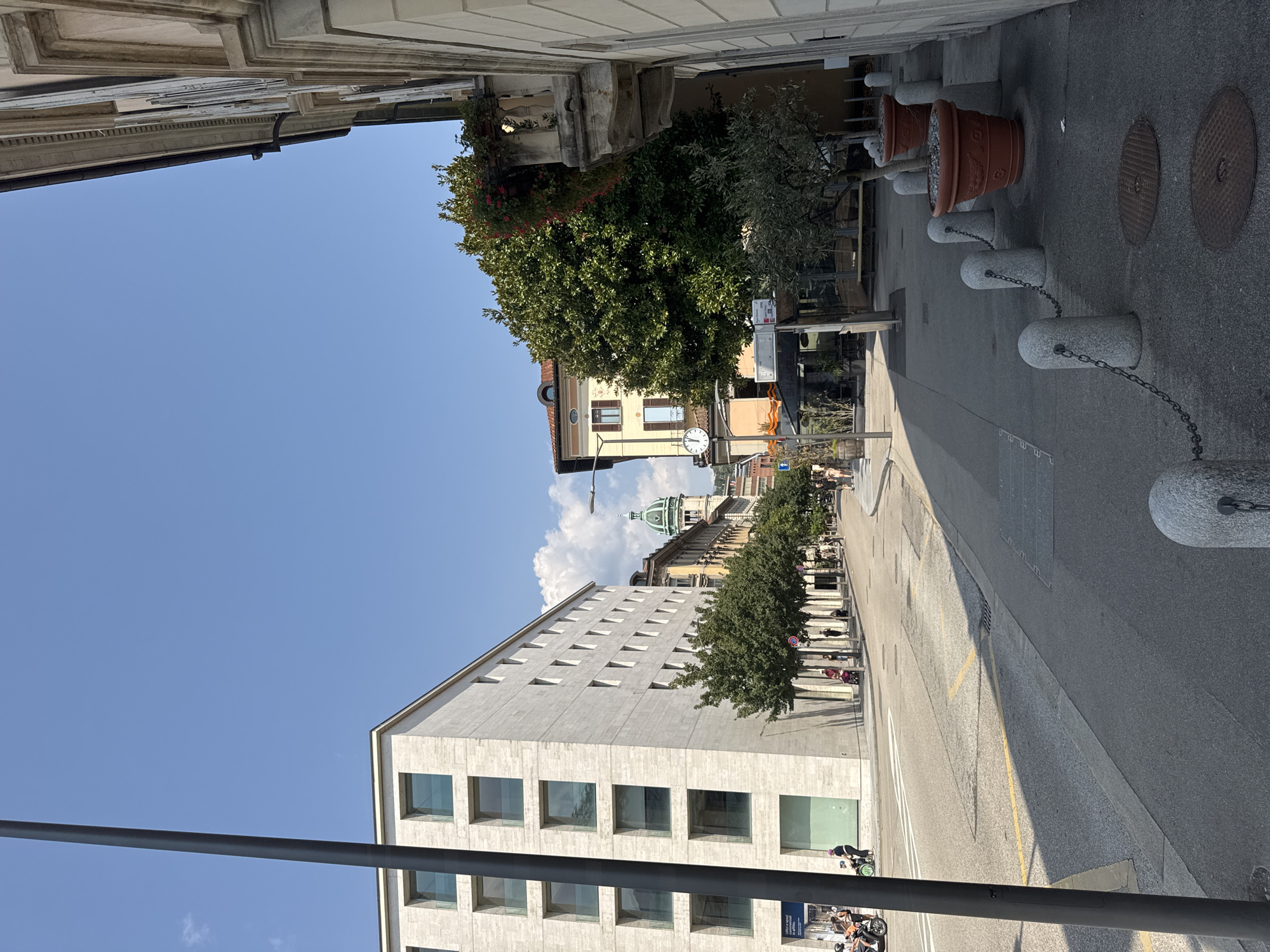 The image shows a street scene in Lugano, Switzerland, under a clear blue sky. On the left is a modern, light-colored building with multiple floors and square windows. In front of this building, a street runs diagonally into the distance. On the right side of the street is a sidewalk with several large, terracotta planters containing trees. A balcony extends from a building on the far right, adorned with flowers. In the background, a domed building is visible, characteristic of city architecture. Several people are present on the sidewalk and street, some riding scooters or bicycles, others walking. A street clock is mounted on a pole near the center of the frame. The overall environment appears to be an urban setting with a mix of modern and older architecture.