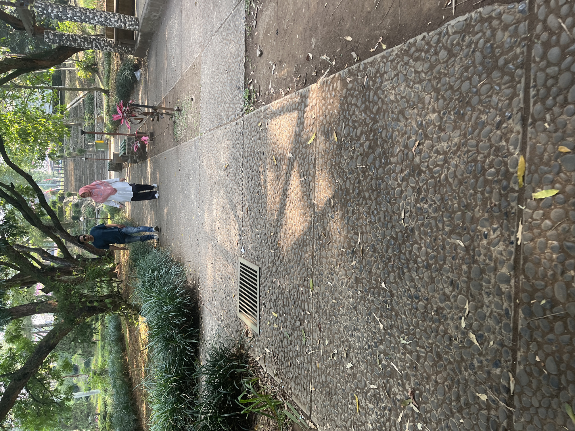 A full shot captures two individuals walking along a pebble-paved path in a park, under the dappled sunlight filtering through a canopy of trees. On the left, a man in a dark blue t-shirt and jeans, wearing sunglasses, walks with his hands in his pockets. To his right and slightly ahead, a woman in a white long-sleeved shirt, dark pants, and a pink hijab walks with her head slightly bowed. The path is bordered by lush green grass on the left and a dirt area on the right. Further in the background, a stone stairway ascends the slope of the park, flanked by stone pillars and benches, suggesting it's within a public urban park environment in Bandung City, Indonesia.