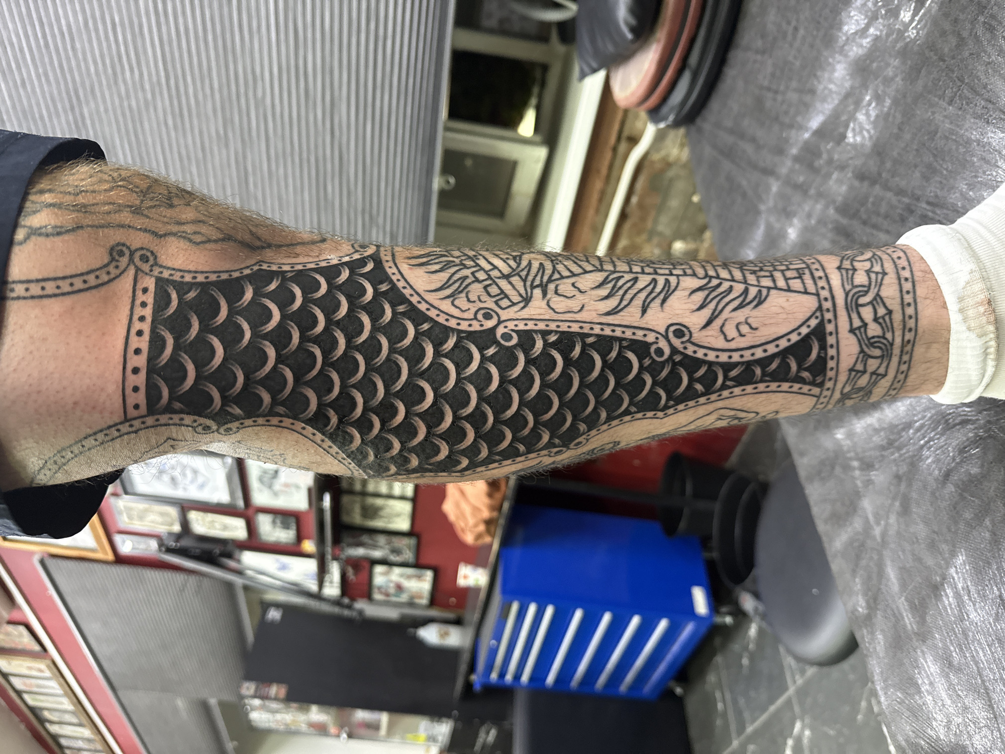 The image depicts a close-up view of a tattoo on a person's lower leg, styled to resemble a piece of armor with scale-like patterns and decorative motifs. The tattoo is predominantly black ink, with some lighter shading visible on the skin around it. The background suggests a tattoo studio environment, featuring a red wall adorned with framed artwork, a blue rolling tool chest, and various other studio equipment like black bins and a piece of furniture covered in plastic sheeting. A window with blinds is visible in the background, showing an outdoor scene that is out of focus. The lighting appears to be artificial, casting shadows and highlighting the texture of the skin and the tattoo ink. The image is a static, close-up shot, focusing on the details of the tattoo and its immediate surroundings. The location is stated to be Moscow, Russia.