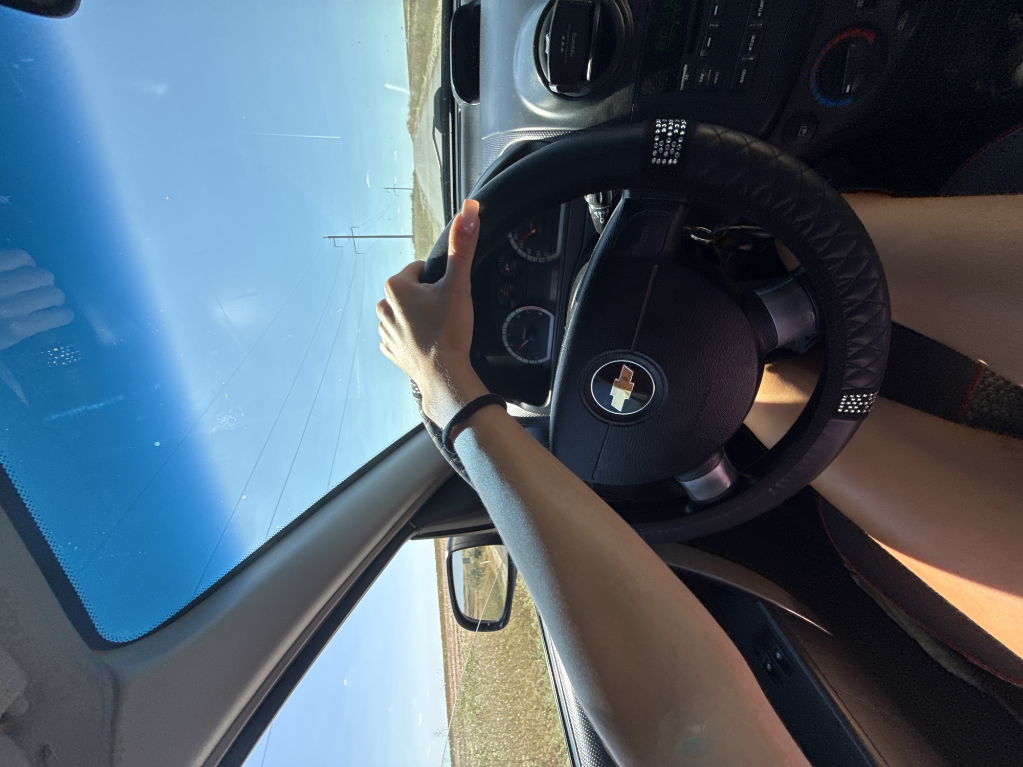A first-person perspective shot from inside a car shows a person's hands on the steering wheel, driving on a sunny day. The steering wheel is black with a diamond pattern and a Chevrolet emblem. The dashboard has visible gauges and controls. Outside the car, the landscape is rural, with fields and power lines visible under a clear blue sky. The location is identified as Cioc-Maidan, Moldova.