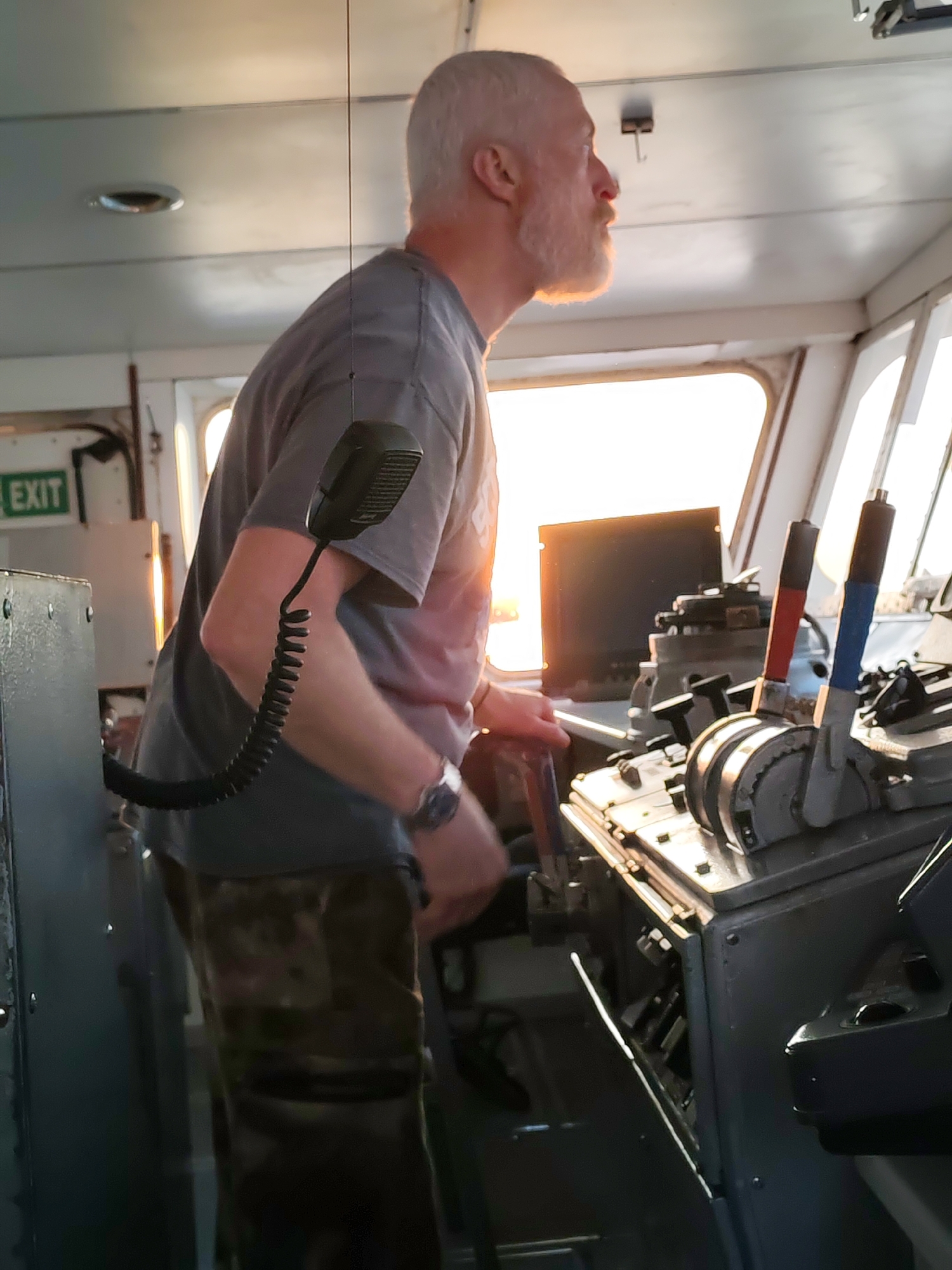 The image depicts a man in the wheelhouse of a ship, likely at sea given the presence of nautical controls and lack of discernible land features through the windows. The man has short, gray hair and a beard. He is wearing a gray t-shirt and camouflage pants. He appears to be operating the ship, his hands are on controls. A coiled microphone is attached to a panel near him. The setting sun shines through the front windows. The location and city are unknown.