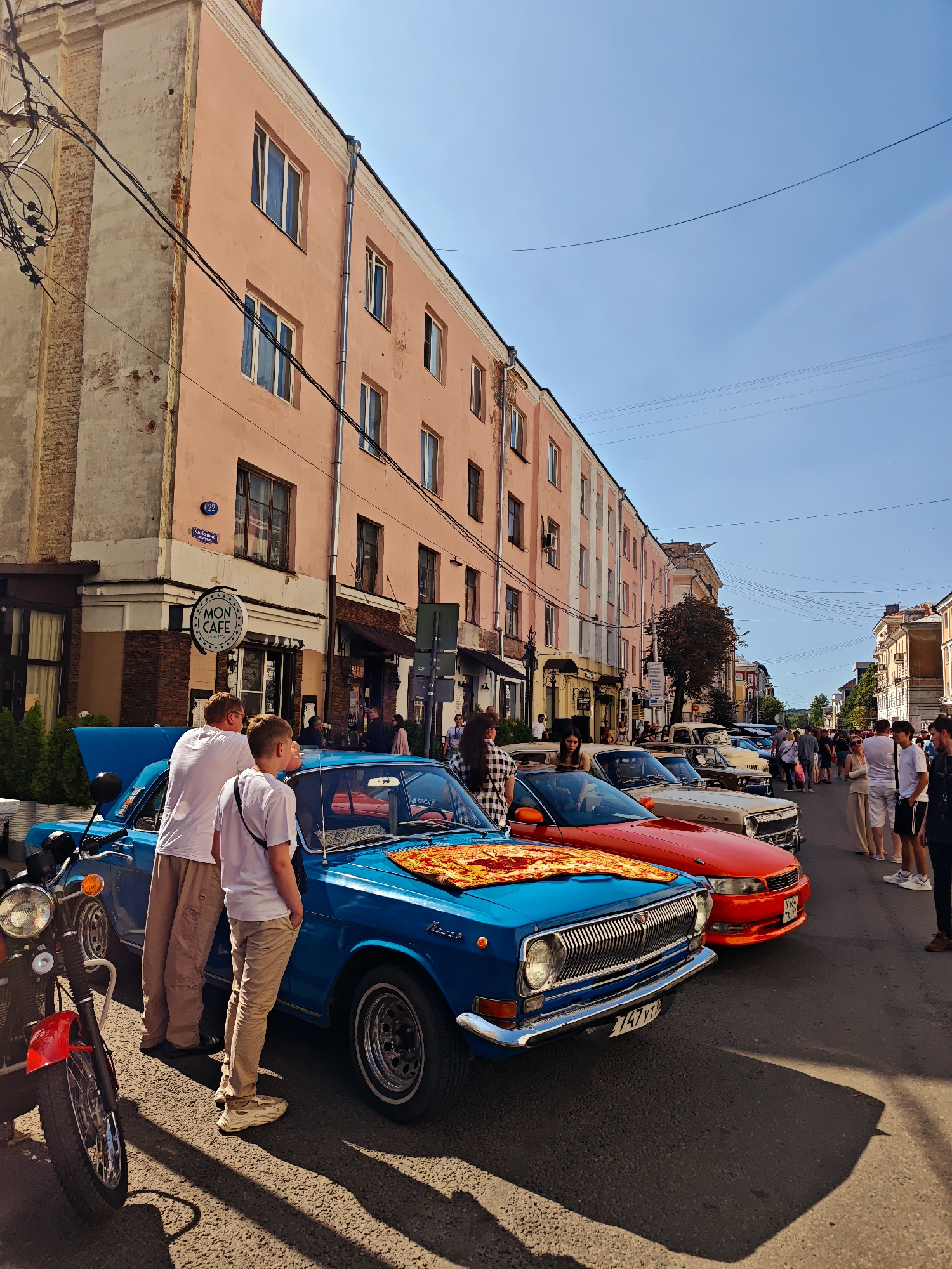 This is a sunny, outdoor, street-level, full-frame, eye-level shot of an urban scene in Tver, Russia, likely during a car show or exhibition. In the foreground, a bright blue vintage car is parked, its hood open. To the left, a red motorcycle is parked. Two men are standing next to the blue car, looking towards it. Several other vintage cars are parked behind the blue car, including a red one and a light-colored one. A crowd of people, both men and women, are dispersed along the street, some looking at the cars and others interacting. The buildings lining the street are two to three stories tall, with pink and cream-colored facades. Overhead, electrical wires are strung across the sky. The street surface is asphalt. A sign for "MON CAFE" is visible on the side of one of the buildings. The sky is clear and blue.