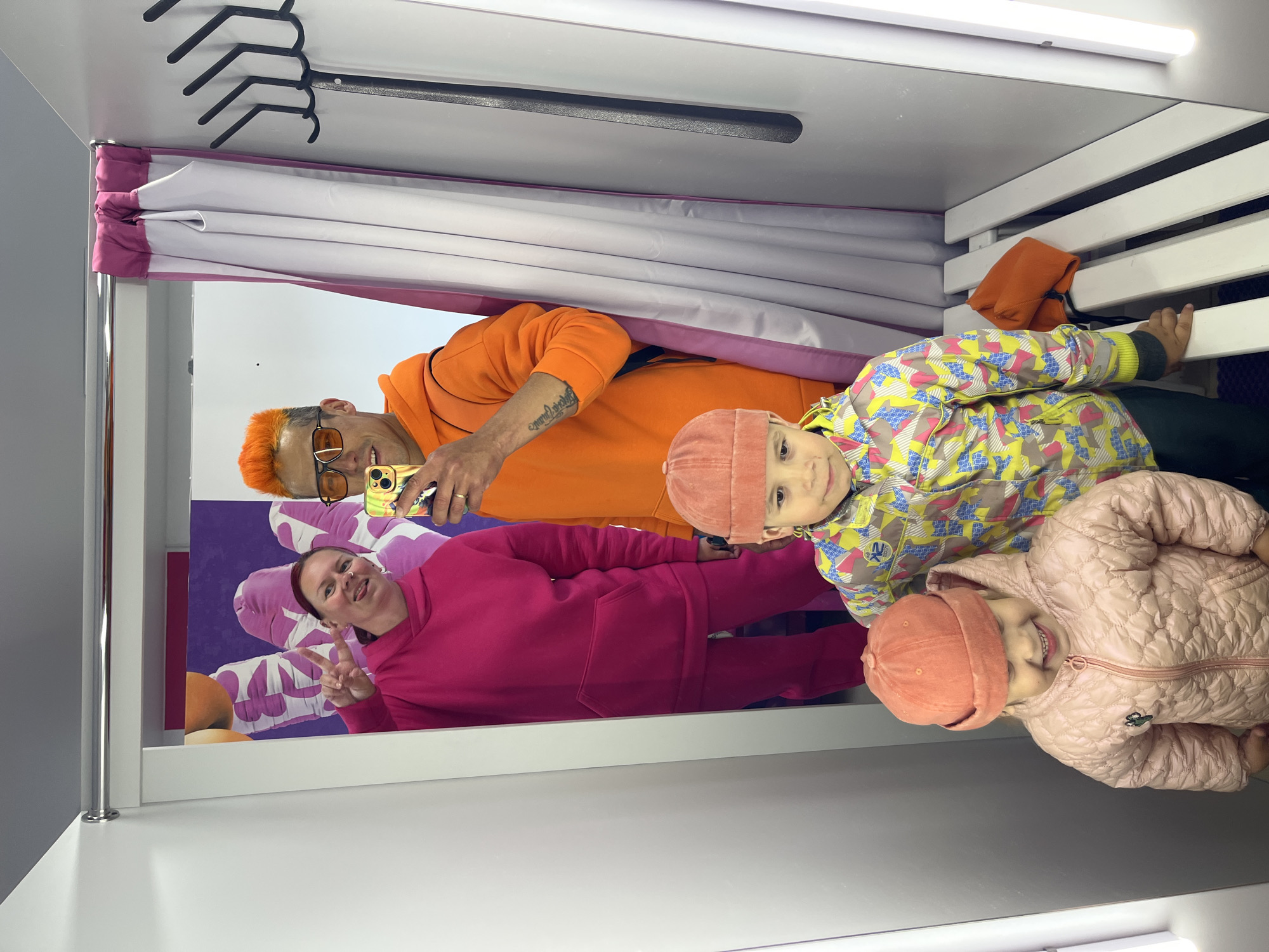 The image shows a group of people in a changing room. There are two adults and two children. The adults are wearing bright colored hoodies, one pink and one orange. One of the adults is holding a phone and taking a picture. The children are wearing jackets and hats. The changing room has a curtain and a rack for clothes. It is unclear from the image whether the location is in the city of Chaikovsky, Russia.