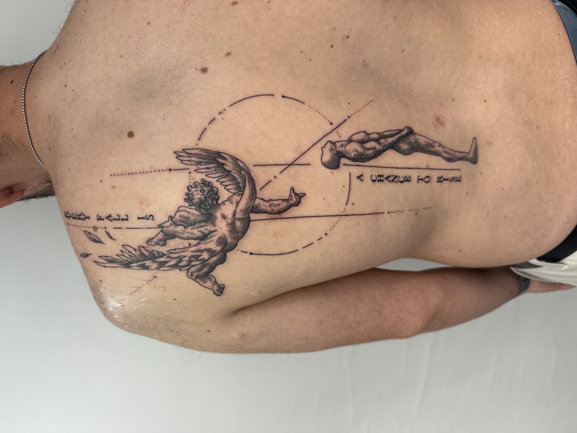 The image captures a detailed black and grey tattoo on a person's upper and mid-back. The skin is light-toned, featuring several moles and fine light hair, with a silver chain visible around the neck on the left. The person is positioned with their back exposed against a plain white background, suggesting an indoor setting.
The tattoo is an intricate design combining classical figures with geometric and blueprint-like elements. On the left, a winged cherub-like figure, rendered with detailed shading, appears in dynamic motion. Below it, the vertically oriented text "EVERY FALL IS" is partially visible. Connecting the figures are dashed lines, solid lines, and arcs that form a diagrammatic backdrop. On the right, a reclining, nude human figure is positioned horizontally and facing upwards. Beneath this figure, the text "A CHANCE TO RISE" is clearly legible. Geometric lines, including a large dashed circle and intersecting straight lines, further integrate the figures thematically. Slight redness around the tattoo suggests it may be recent or healing. The overall artwork portrays a symbolic narrative of falling and rising. The provided location context for this image is Sofia, Bulgaria.