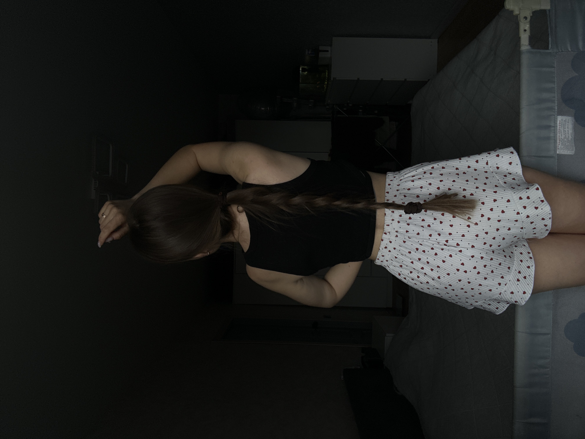 A person, presumably female, is lying down in a dimly lit room. She is wearing a black tank top and a white, short skirt with red heart patterns. Her hair is long and braided. The person is lying next to a bed with a gray patterned duvet. A dresser is partially visible in the background. The location cannot be determined from the provided image.