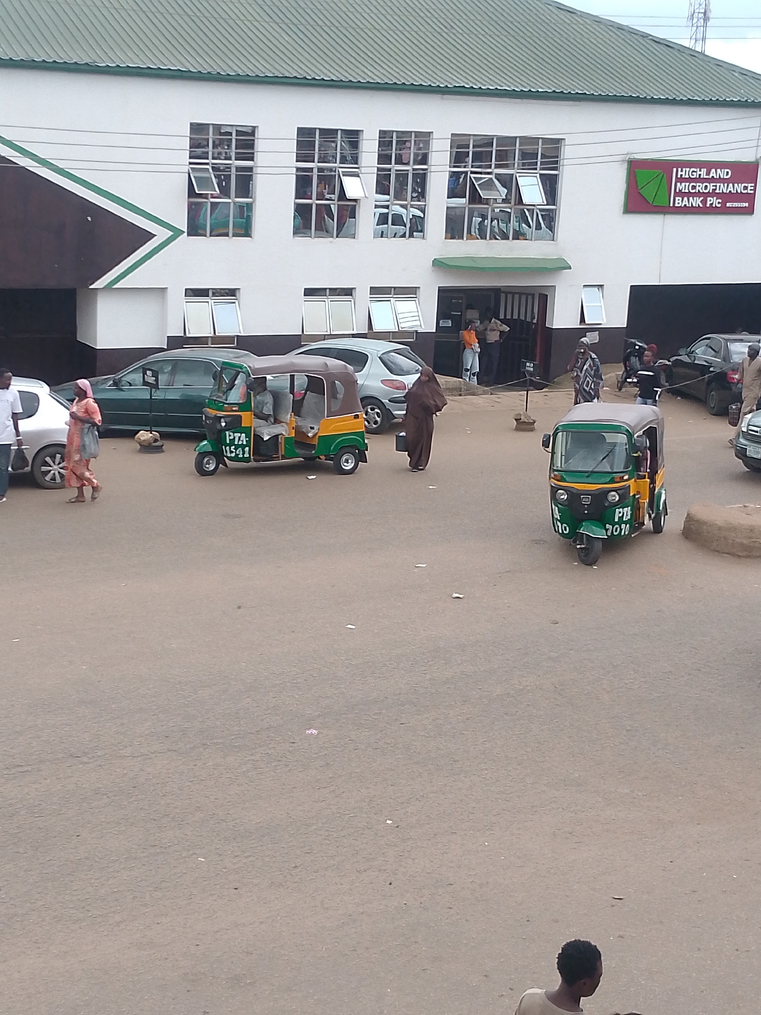 The image depicts the exterior of Highland Microfinance Bank Plc in Bukuru, Nigeria. The building is a two-story structure with a green, corrugated metal roof. The front of the building is white with dark brown accents and a large green chevron design on the left side. There are numerous windows on the upper floor, many of which are open. A prominent sign on the right side reads "HIGHLAND MICROFINANCE BANK PIC".
In front of the bank, there is a paved area where several vehicles and people are present. Two auto-rickshaws, commonly known as "keke napep" in Nigeria, are parked. One is green and yellow with "PTA 11541" visible on the front. The other, closer to the foreground, is also green and yellow with "PTA 7070" on its front. Several cars are also parked in the area, including a dark-colored sedan to the right and a green sedan to the left, in front of which a woman in an orange and pink patterned dress and a light pink headscarf is walking. Another woman in a dark brown hijab is walking away from the camera towards the left.
Several people are visible around the entrance of the bank. Two men and a woman are standing near the entrance doors. Further to the right, a woman in a patterned blue and white garment is walking, and a man in a dark shirt and jeans is standing near a motorcycle. The scene appears to be set during daylight hours, with overcast skies suggesting cloudy weather. The overall impression is one of a busy, commercial setting.