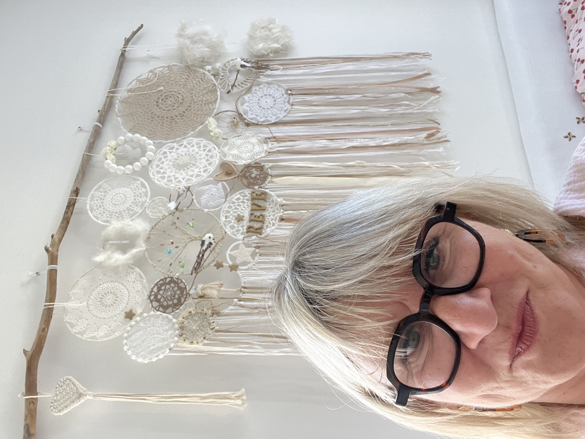 The image presents a selfie taken indoors, likely within a residential setting. The subject is a woman with fair skin, wearing black-rimmed glasses and exhibiting gray/blonde hair. She appears to be holding a decorated branch or wall hanging.
The decoration is attached to a branch and features an assortment of handcrafted elements, including crocheted circles, fabric strips, beads, and small decorative items. The colors are predominantly white and cream. There is the word "REVE" written.
The location is Montélimar, France.