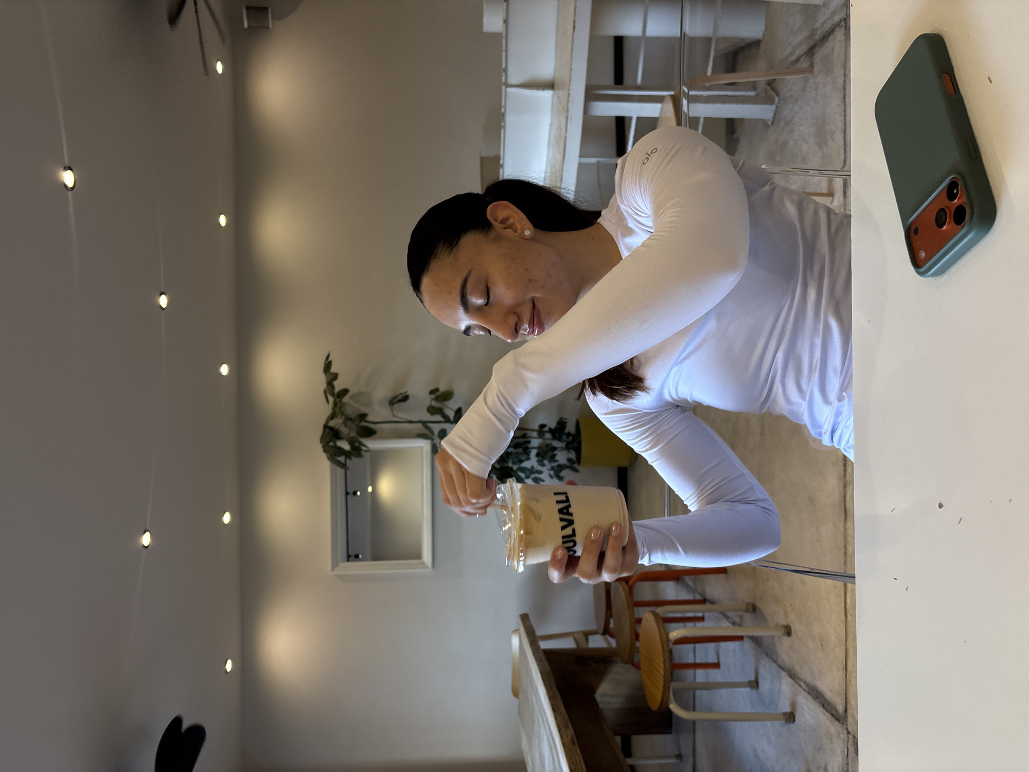 A woman in a white long-sleeved shirt is holding a clear plastic cup labeled "JULVALI." She is positioned at a table in what appears to be a café or restaurant. The background shows a wall with several small, circular lights, plants, a mirror, and chairs. A cell phone rests on the table. Based on the "JULVALI" branding on the cup, the location is likely in the city of Puebla, Mexico.