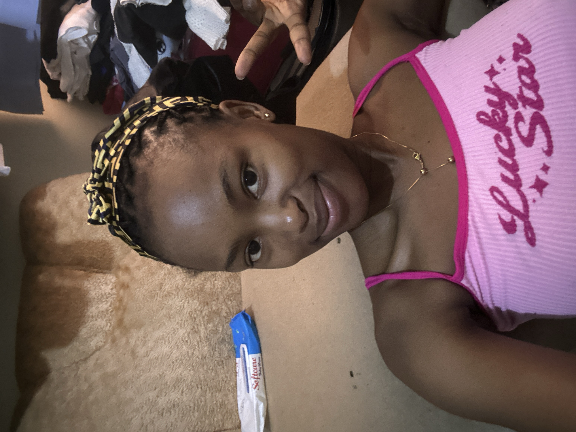 A young Black woman with braided hair and a patterned headscarf is posing for a selfie on a sofa. She is wearing a pink ribbed tank top with "Lucky Star" written on it in pink lettering. She has a gold necklace with two charms. To her left is a package of "Softcare Baby Wipes". In the background, to her right, are piles of clothes. The setting appears to be indoors, likely a living room in Jos, Nigeria, given the provided context. The woman is smiling and making a peace sign with her right hand.