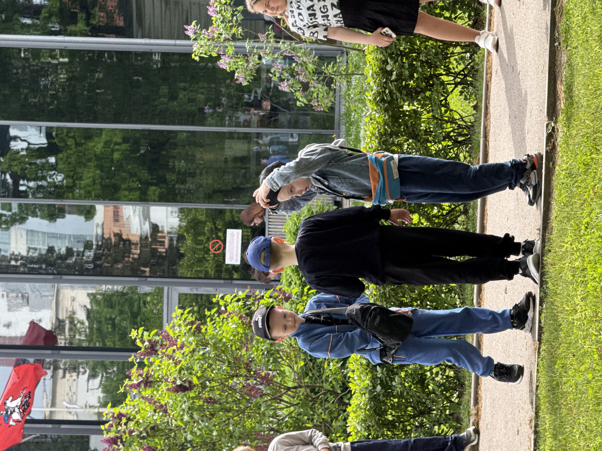 A group of children are standing on a sidewalk alongside a building with mirrored windows. Lilac bushes are in bloom nearby. The location is likely Moscow, Russia, due to the presence of a Moscow coat of arms flag attached to the building. One child wears a black Puma cap, another a blue baseball cap, and a third a denim jacket. The children appear to be students on an outing or field trip.
