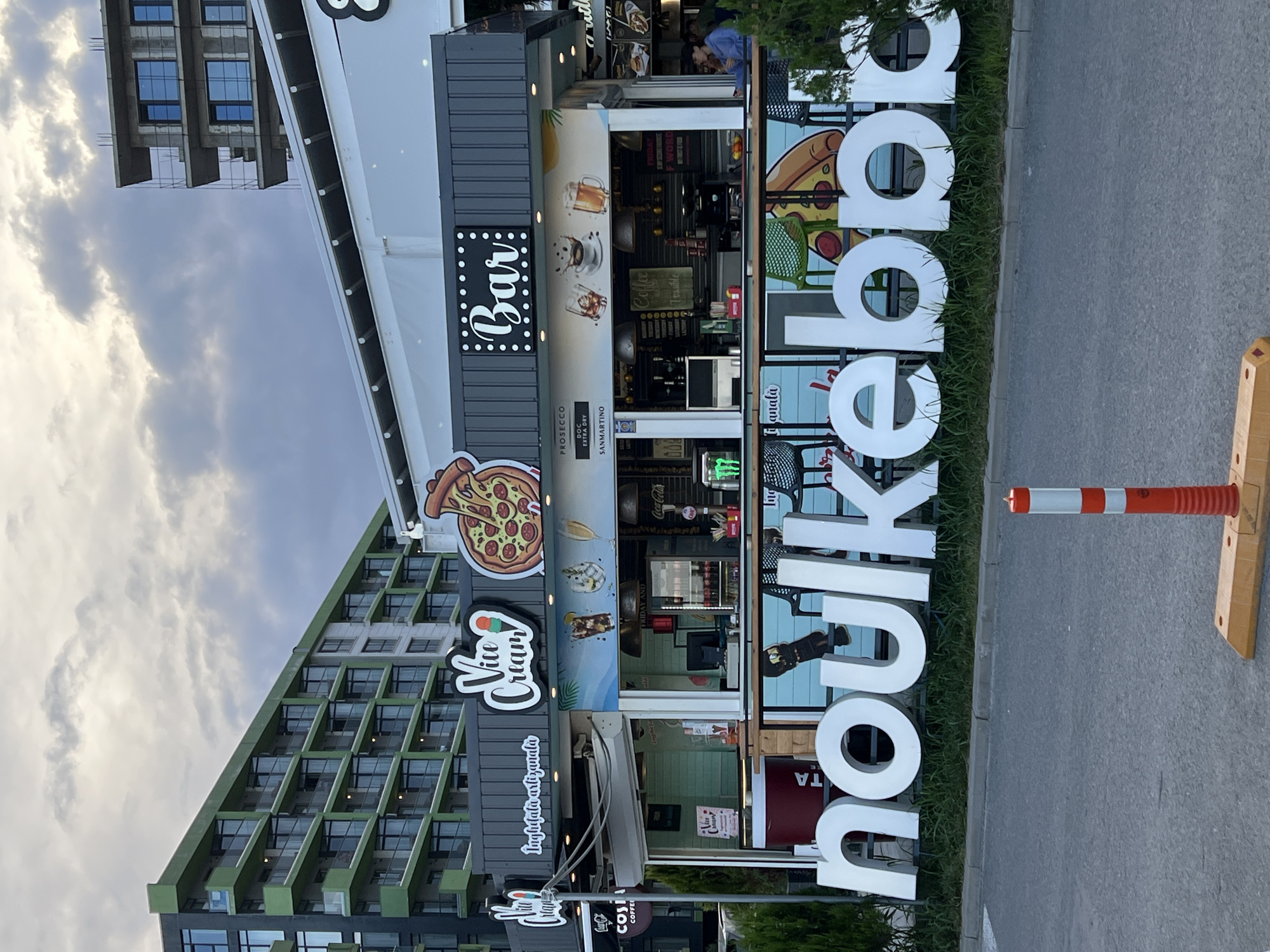 The image depicts a storefront featuring "Vito Cream" and "Bar" signage, with a large white sign in the foreground reading "noulkebbp". The storefront has a modern design with colorful graphics advertising pizza, ice cream, and drinks. A multi-story building with green balconies is visible in the background, along with a building under construction. The scene appears to be in a commercial area, possibly along a promenade or street in Năvodari, Romania. There are no visible people interacting with the storefront in the foreground, but a person is seen inside the bar area in the background. The overall atmosphere is casual and commercial, with an emphasis on food and beverage services.
