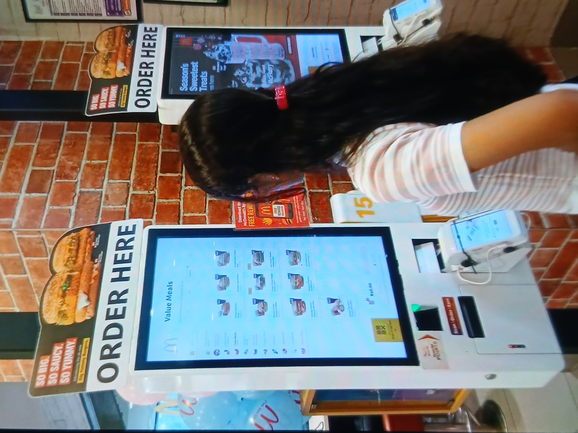 In Setia Bakti, Indonesia, a young woman with long dark hair and wearing a striped shirt stands in front of a McDonald's self-order kiosk. She is looking at the screen, which displays a menu with various "Value Meals." Another self-order kiosk is visible to the right, featuring a "Season's Sweetest Treats" advertisement. The kiosks are positioned against a brick wall. The image captures a typical scene of a customer interacting with technology for food ordering.