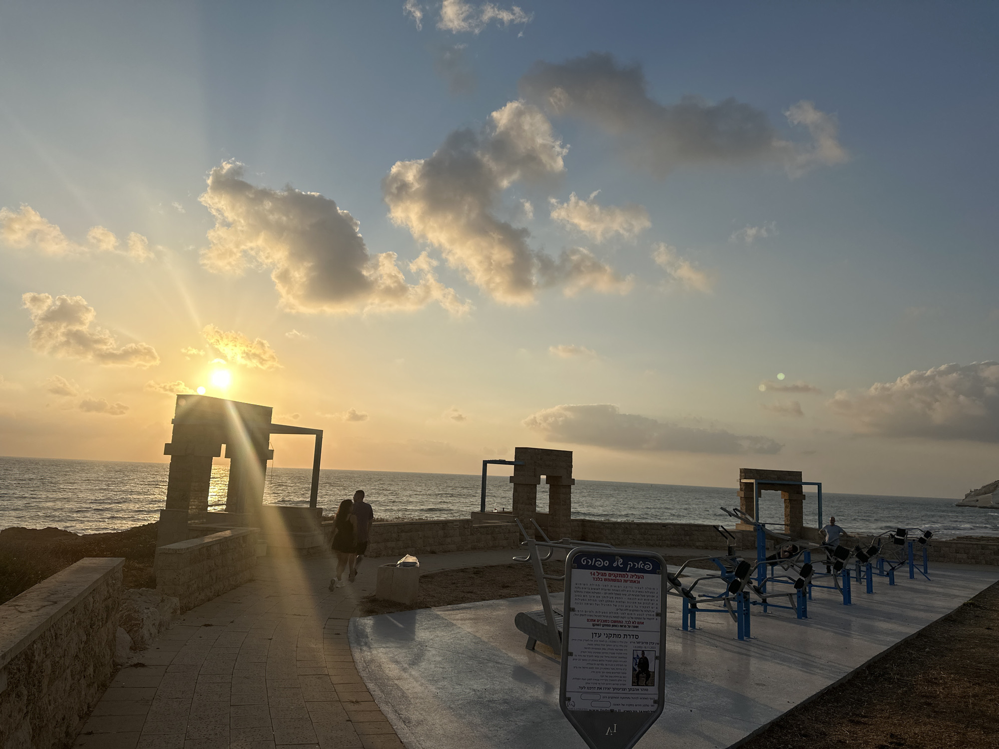 This image captures an outdoor exercise area near the coast in the Mate Asher Regional Council, Israel, at sunset. The sun, low on the horizon, casts a warm, golden glow across the scene, with visible sun rays piercing through scattered clouds. The sea stretches out to the left, with gentle waves lapping against a rocky shoreline and a stone-built structure with an arched opening.
In the foreground, a paved walkway curves around an area equipped with various exercise machines, primarily in blue and silver. A signpost with text in Hebrew, likely providing instructions or information about the exercise equipment and local area, stands prominently in the lower right.
Two individuals are visible walking away from the camera on the paved path. One person is wearing a dark t-shirt and shorts, and the other is in a dark dress. Further back, near the arched structure, another person appears to be using one of the exercise machines. The ground surrounding the exercise equipment is covered in dry grass and dirt. The overall scene conveys a sense of a public recreational space in a coastal setting during the evening.