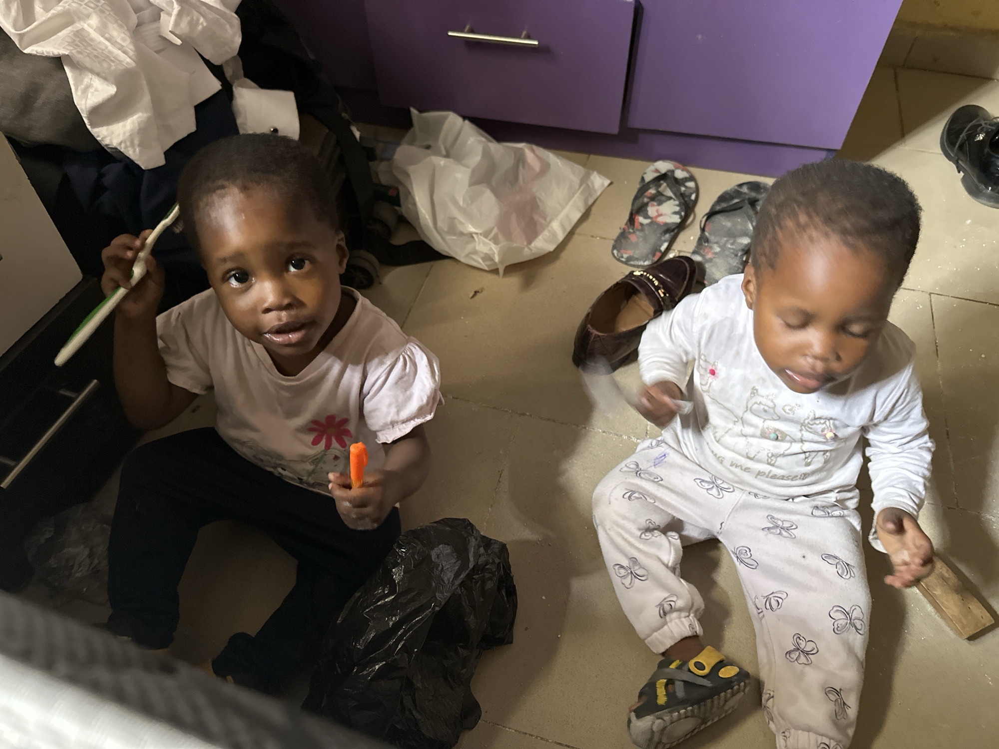 In Narayi, Nigeria, two young children are on the floor, seemingly playing amongst discarded items. The child on the left, wearing dark pants and a white t-shirt with a pink flower graphic, holds an orange object and a toothbrush, looking towards the camera. The child on the right, dressed in white pajamas with butterfly prints, lies on their back with their eyes closed. The environment appears to be a cluttered area with items such as a black trash bag, a white plastic bag, a brown loafer, a pair of flip-flops, and a purple cabinet.