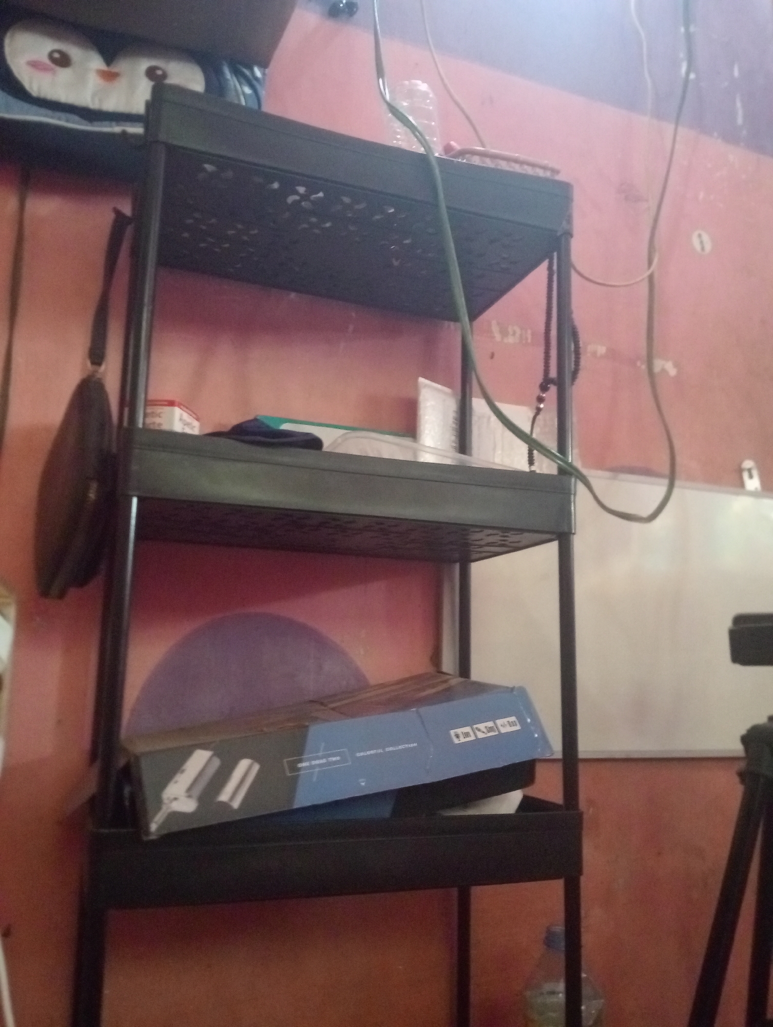 The image depicts an indoor scene, likely a room in Bloro, Indonesia, featuring a black, multi-tiered shelving unit against a reddish-orange wall. The top shelf holds a blue bag with cartoon penguin faces. The second shelf contains a small white box with red lettering, a dark blue object, and a white box with text. A dark brown, oval-shaped bag hangs from the left side of the shelving unit. The third shelf has a blue and white box with text and graphics, resting on a white object. The bottom of the shelving unit is empty. A clear plastic bottle with a blue cap is visible on the floor near the bottom of the shelving unit. Electrical wires and cables are draped across the scene. A whiteboard is partially visible on the right side of the wall, and a black tripod is also present in the background. The lighting suggests an indoor environment, possibly with ambient light from a window or overhead source.