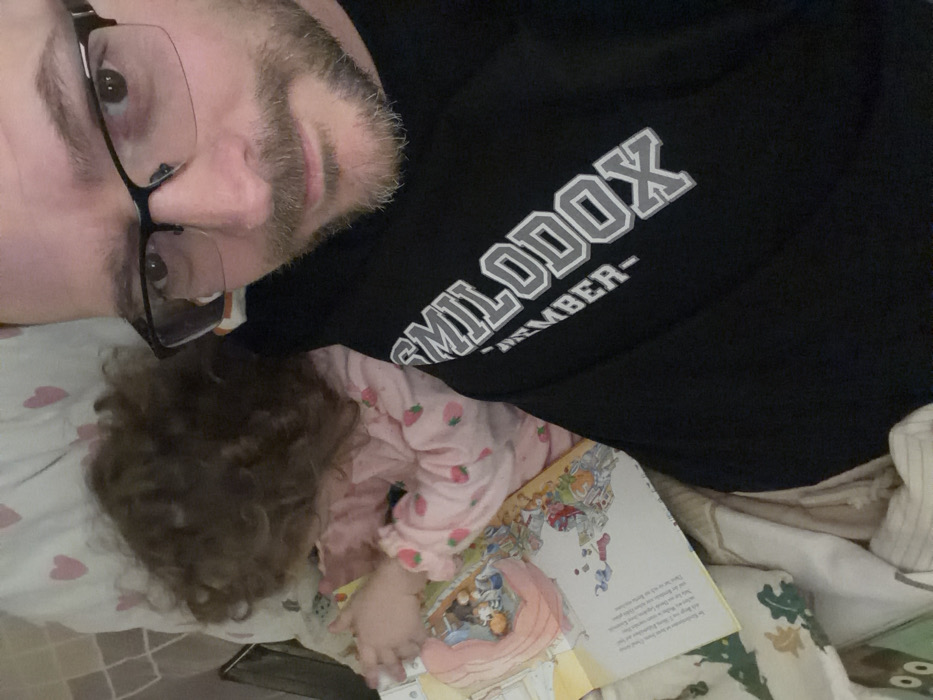 An adult male and a young child are present in an indoor setting in Trassem, Germany, likely a bedroom. The adult male, with short brown hair, a beard, and black-framed glasses, occupies the right half of the frame, looking directly at the camera in what appears to be a selfie. He wears a black long-sleeved shirt with the word "SMILODOX" and "-TEMBER-" visible in grey lettering. To his left, a child with curly brown hair is partially visible, lying down. The child is wearing pink pajamas adorned with a strawberry pattern. An open children's book, featuring colorful illustrations and text (including German words), lies between the child and the adult. The child's hands are near the book. Bedding includes a white pillow with pink heart patterns behind the child and other blankets in shades of cream, brown, and green with a patterned design.
