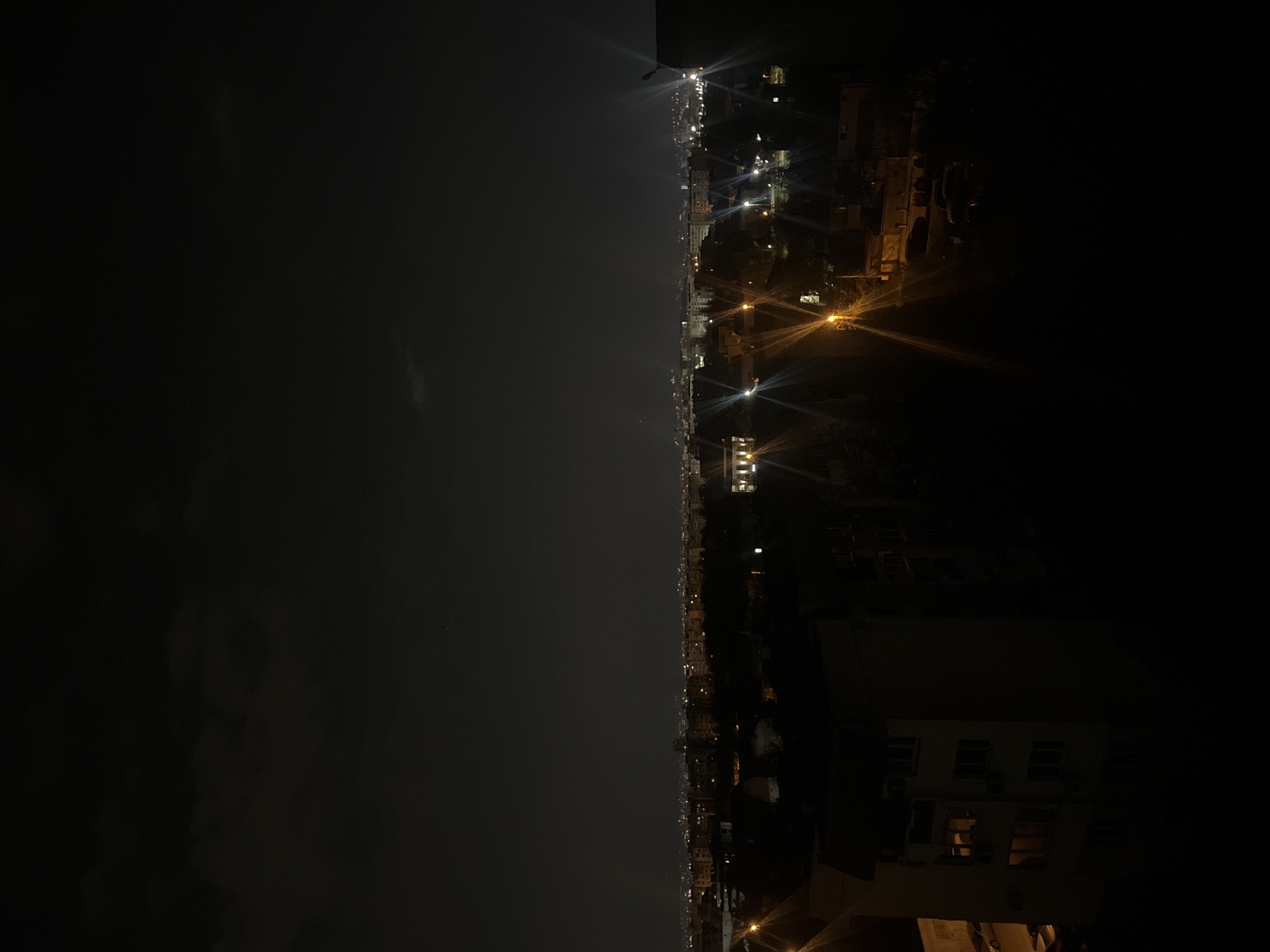 The image depicts a nighttime cityscape, likely in Sofia, Bulgaria, based on the architectural styles. The scene is divided, with the left side dominated by a dark sky and possible cloud cover, while the right side shows a lit urban area with buildings and streetlights. Individual buildings are visible, with their windows illuminated. Streetlights create bright points of light with starburst effects due to the camera lens. The overall atmosphere is dark and slightly hazy. There are no people visible in the image.