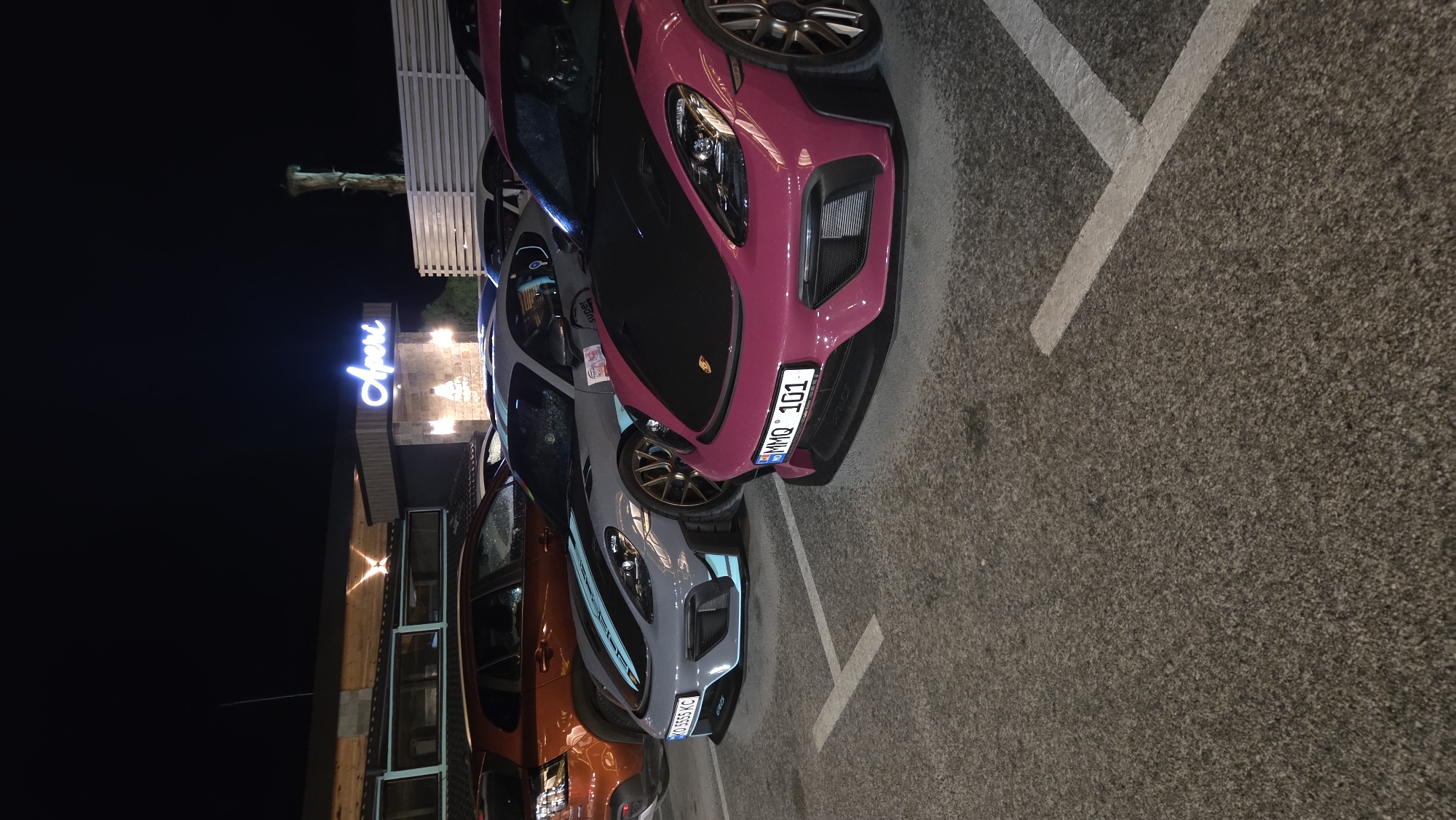 Captured at night, the scene presents a row of parked cars next to a building displaying a neon sign that reads "Aperi". The closest car is painted in a pink hue and has a license plate with the identifier "MMQ 101". Beside it, another car boasts a grey exterior with light blue accents. Further back, an orange vehicle is visible. The location, though unknown, appears to be a city setting with a building offering some sort of dining or social experience, indicated by the "Aperi" sign. The vehicles are parked in designated parking spaces marked by white lines on the asphalt.