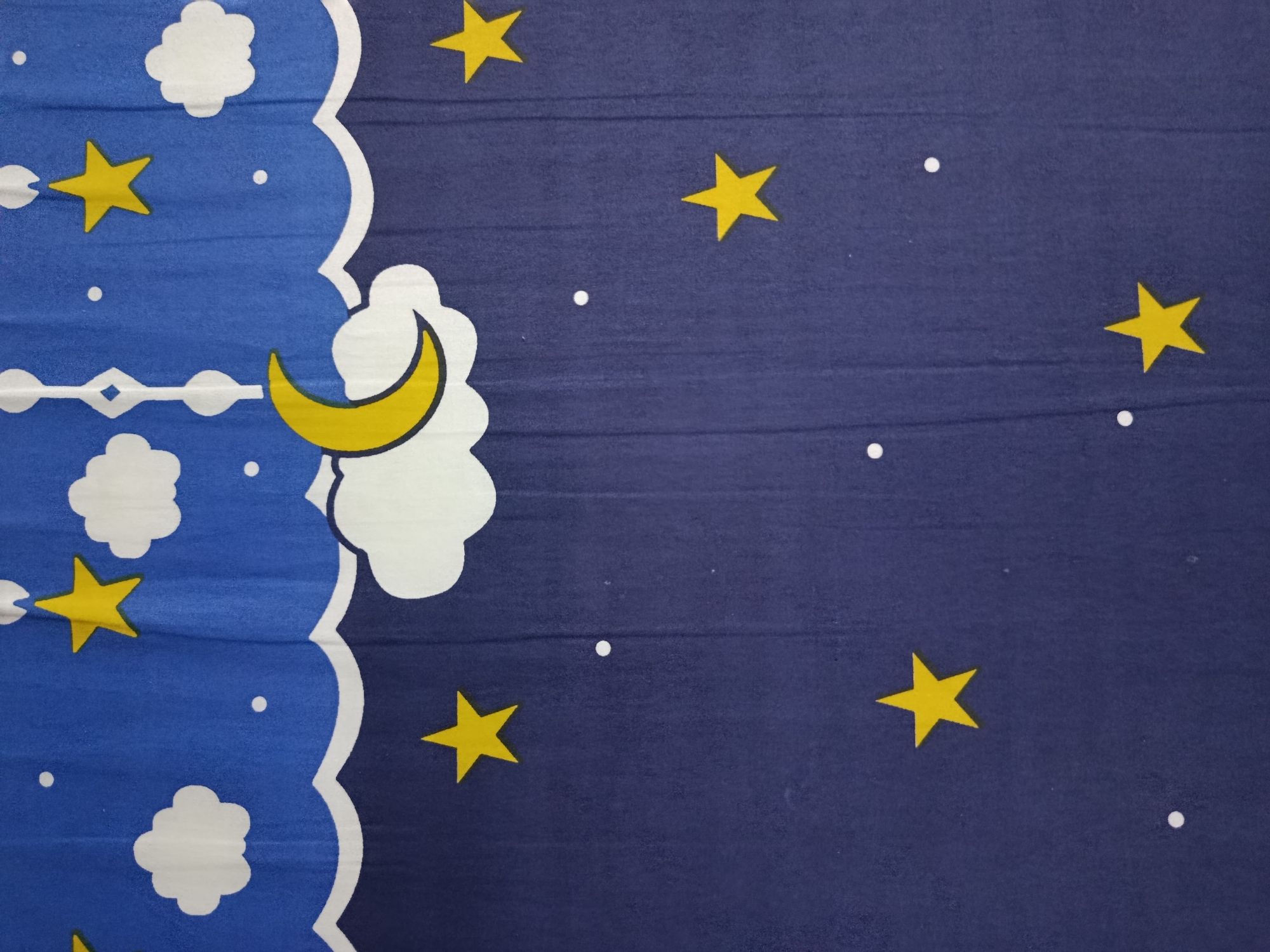 The image displays a close-up view of a fabric with a night sky theme. The fabric is predominantly dark blue, representing the night sky, and is adorned with several yellow stars of varying sizes and white cloud shapes. A crescent moon, also in yellow, is visible on the left side of the image, partially obscured by a white cloud outline. The fabric appears to have a slightly textured surface, possibly due to its material and the way it is illuminated. The pattern is repetitive and suggests it could be part of bedding, a curtain, or another textile item. The specific location in Wanasari, Indonesia, cannot be determined from the visual information presented in the image alone.