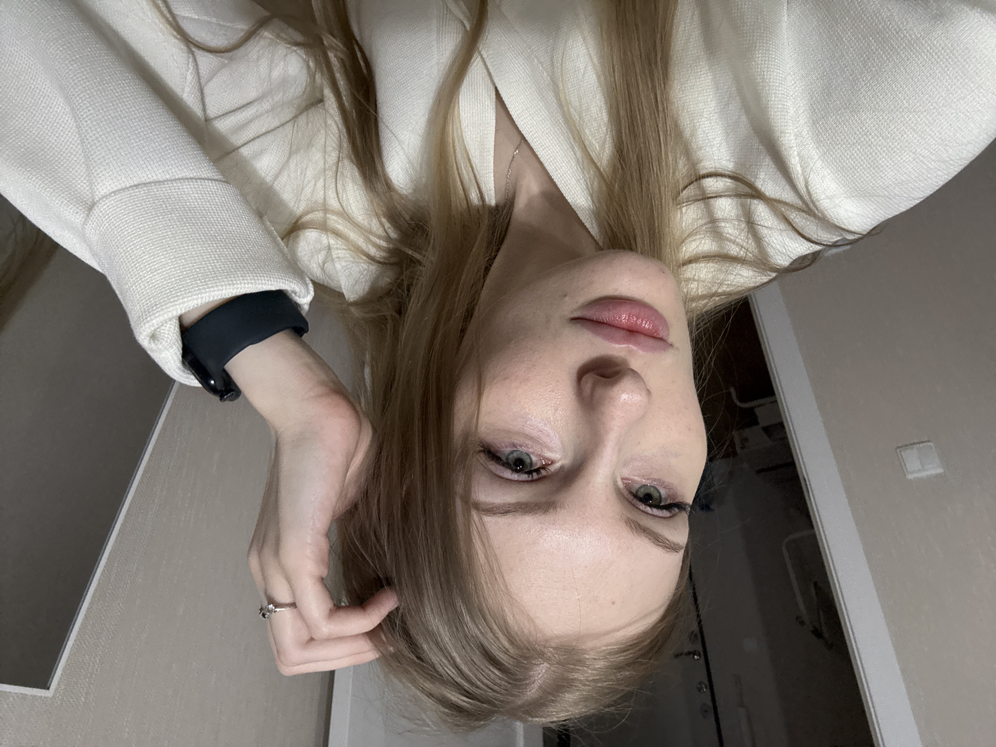 A vertically oriented photograph shows a person with long blonde hair, wearing a white blazer and a black smartwatch. They are looking directly at the camera. A silver ring is visible on one of their fingers. In the background, there is a doorway and a light switch on a wall. It is not possible to confirm the location as Perm, Russia from the provided image.